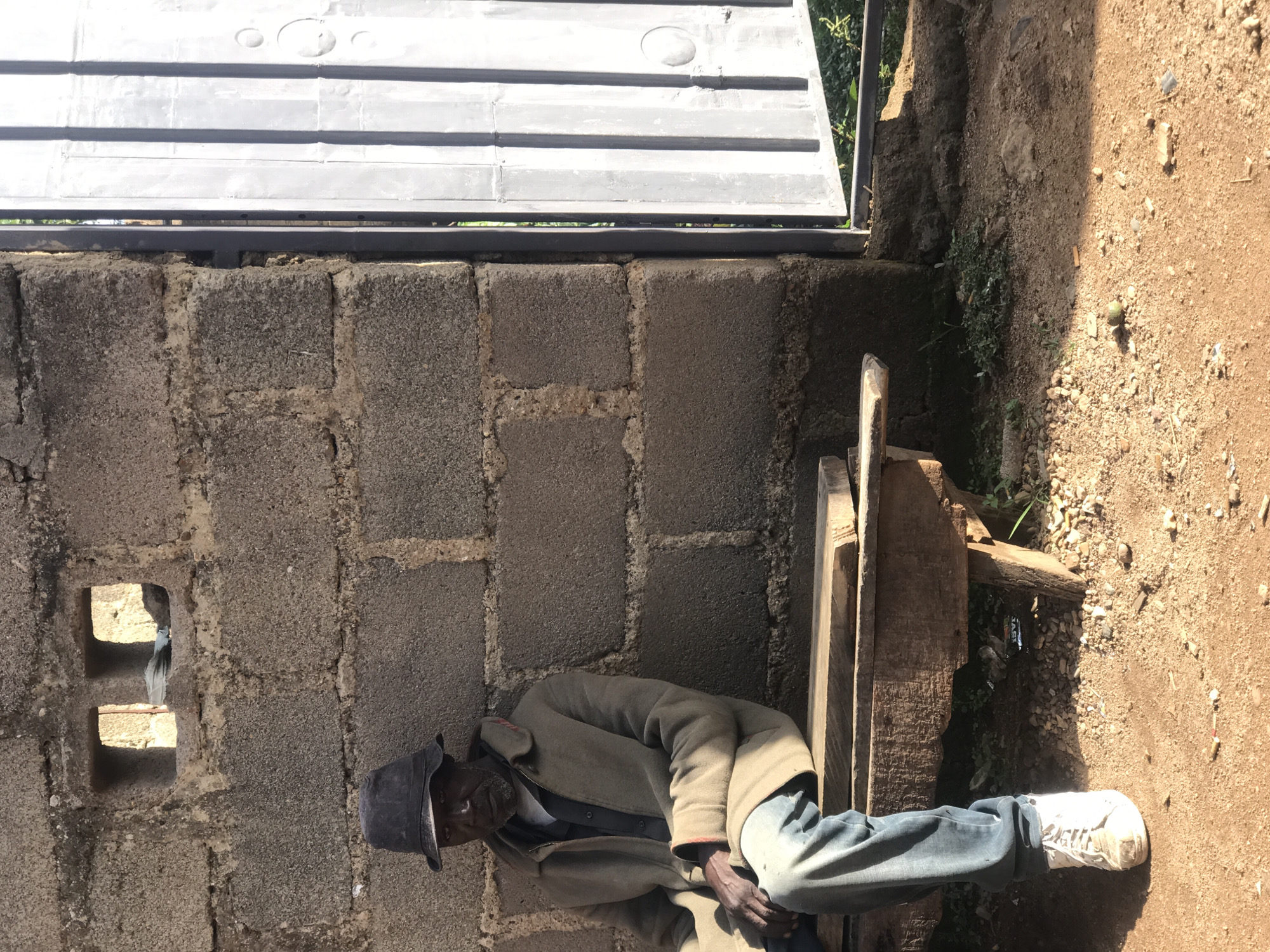 An elderly African man wearing a dark hat, a grey jacket, and blue jeans sits on a wooden bench outside a building made of concrete blocks. The building has two small openings in the wall. To the right of the man is a tall, grey metal gate. The ground is covered in reddish-brown dirt and small stones. The scene appears to be in an outdoor area of Kabong, Nigeria.