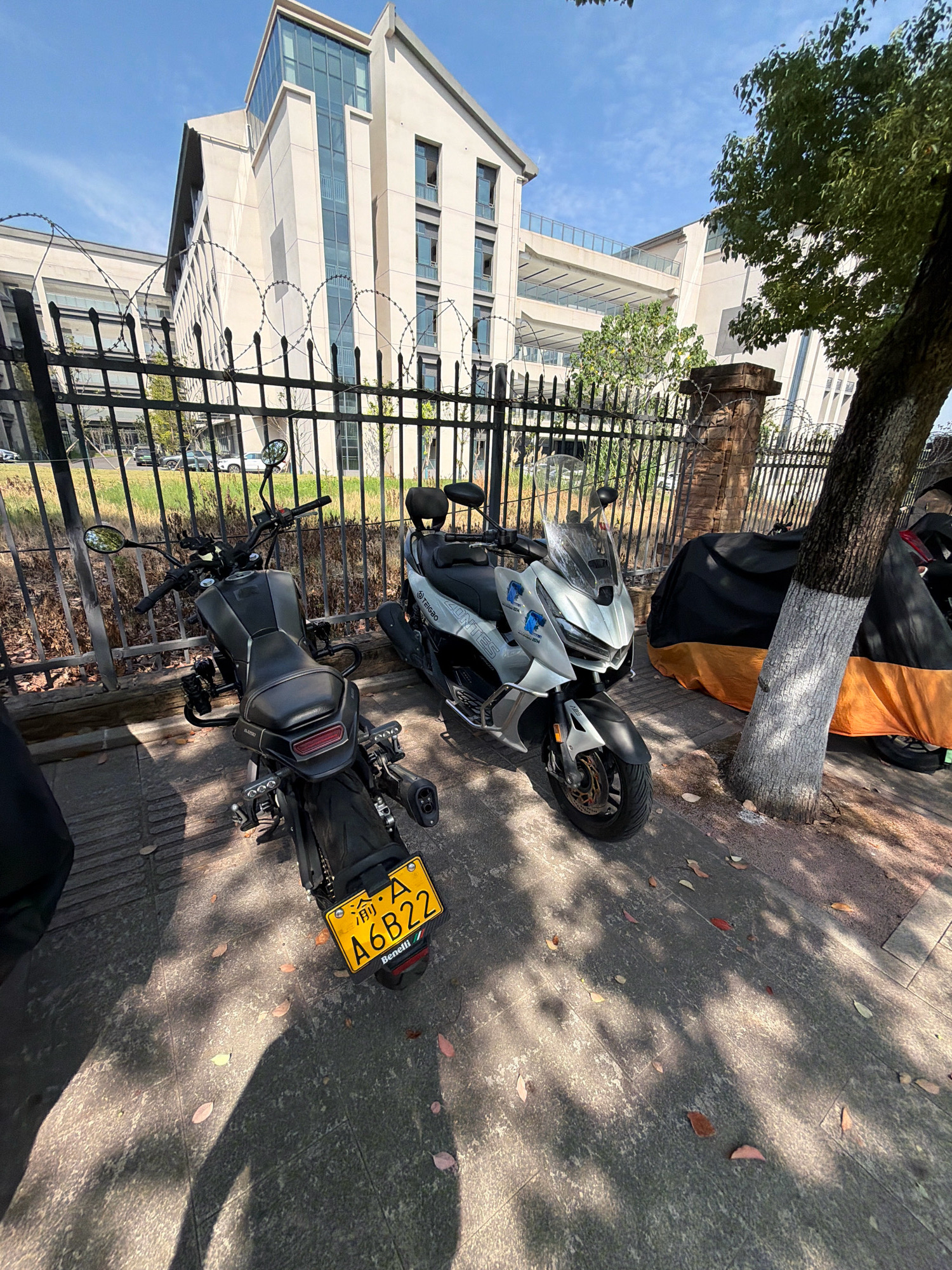 In the city of Jiangjin, China, on a sunny day with a clear blue sky, a street scene unfolds. A black motorcycle with a yellow license plate that reads "A6B22" is parked on a paved walkway. To its right, a silver scooter with a transparent windshield is also parked. Behind the scooters, a metal fence topped with barbed wire separates the walkway from a grassy area. Beyond the fence, a modern, multi-story building with large windows stands prominently. A large tree with dappled sunlight filtering through its leaves is situated to the right of the scooters, casting shadows on the walkway. Another motorcycle, covered with a black and orange tarp, is visible behind the tree. The ground is scattered with fallen leaves, indicating an autumnal or late summer setting. The overall scene depicts a quiet urban environment, possibly near a school or office complex, with parked vehicles on a sunny day.