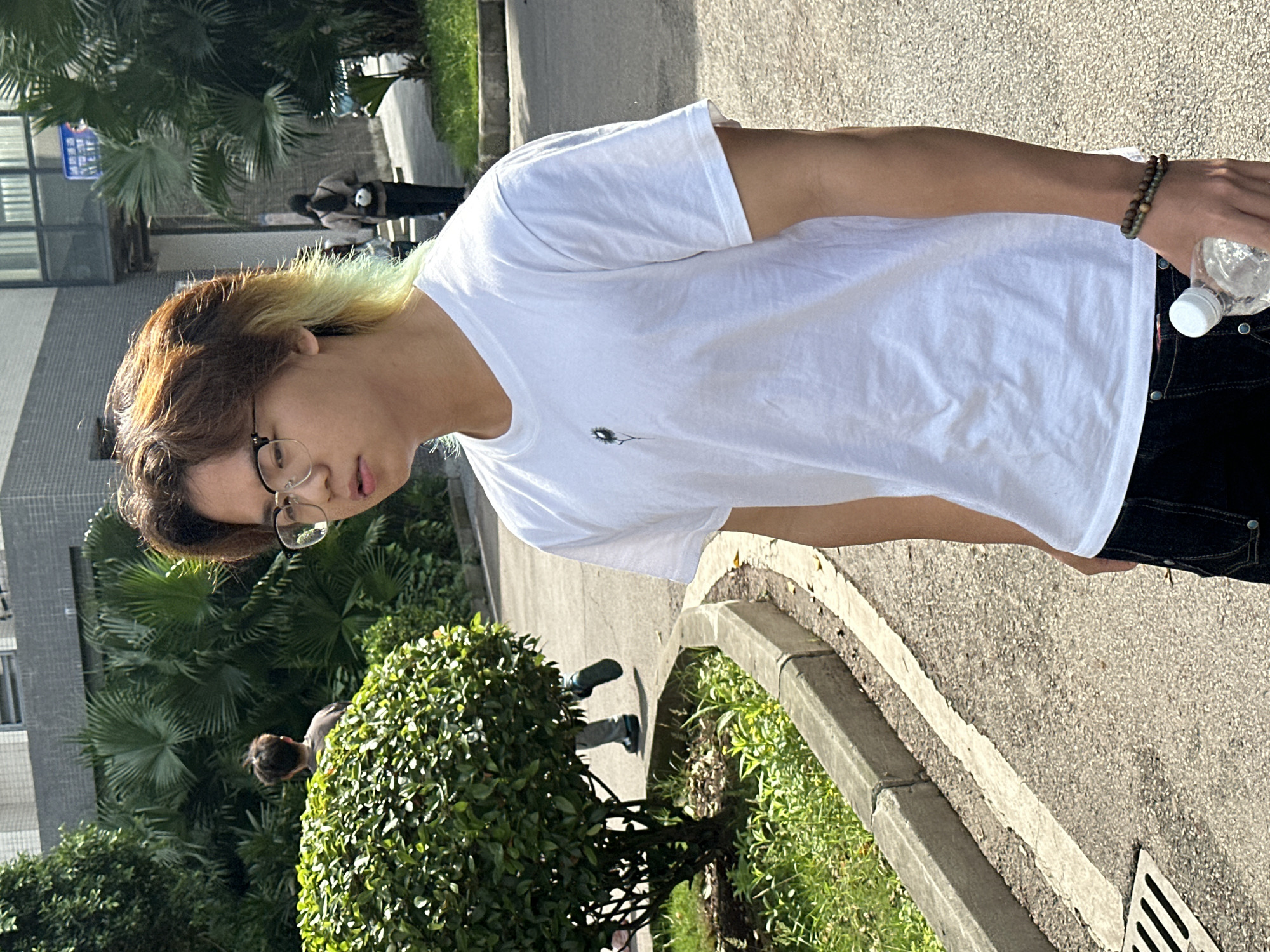 A young individual wearing a white T-shirt and dark jeans stands on a paved sidewalk. They are holding a water bottle. The individual has brown hair with blonde streaks and wears glasses. A bracelet is visible on their wrist. The background features trees, bushes, and a building. Other people are seen walking in the distance. The location is likely Jiangjin, China.