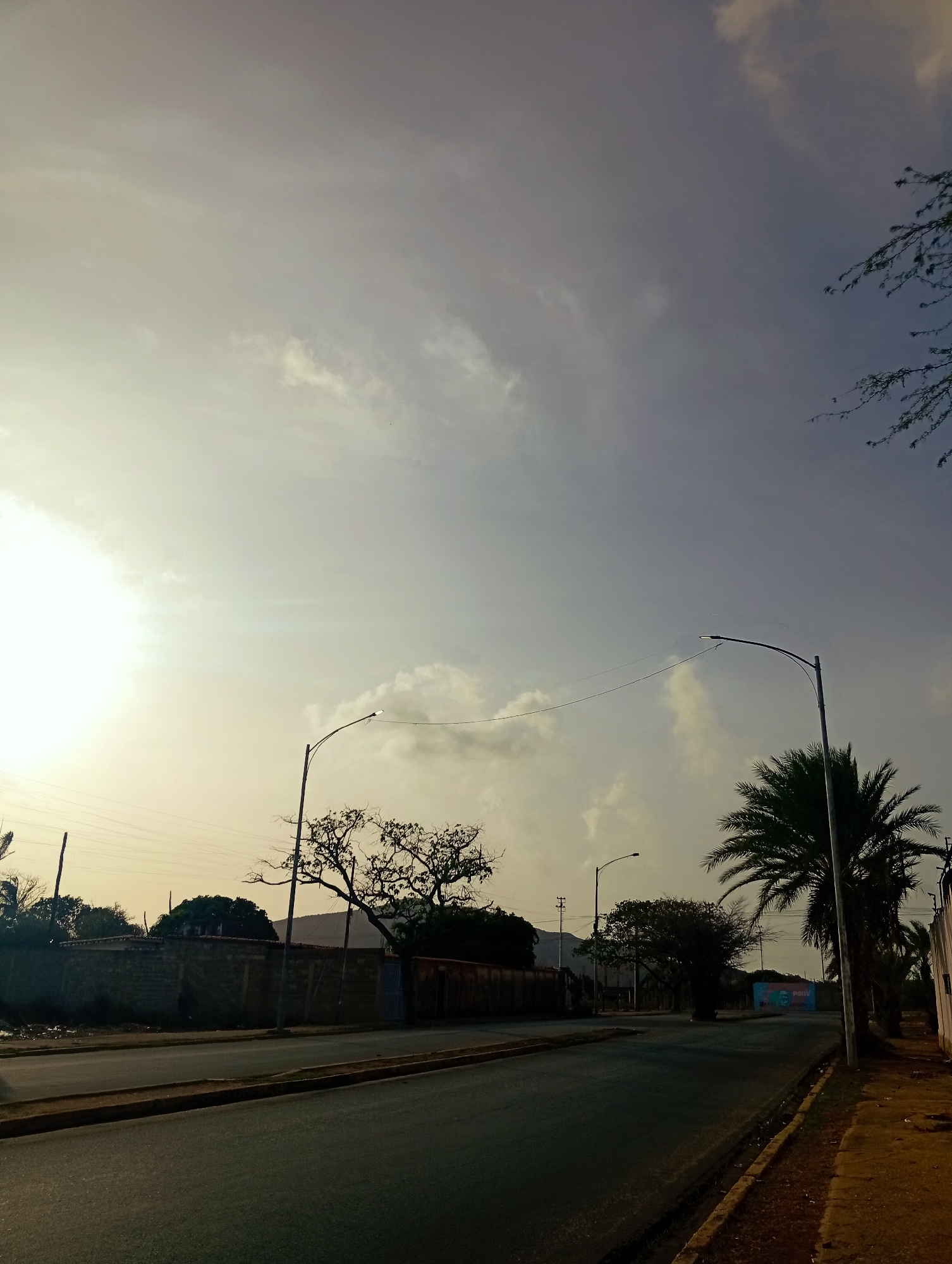 The image depicts a street scene in Juan Griego, Venezuela, likely during late afternoon or early morning given the low angle of the sun and the hazy sky. The foreground shows an asphalt road with a raised sidewalk on the right side. The road appears to be two lanes, with a yellow painted curb. Along the left side of the road, there is a low, brick wall. Beyond the wall are trees and some structures. On the right side of the road, there are several palm trees and a few buildings in the distance. A street lamp is visible in the middle ground. In the far background, mountains can be seen. The sky is partly cloudy and has a yellowish-grey hue. There are no people or vehicles visible in the image.