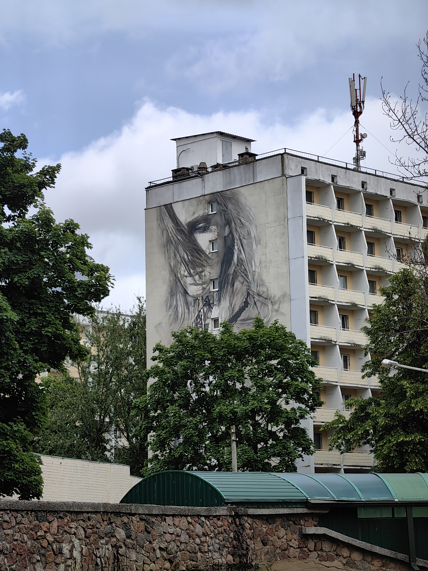 The image depicts a large building in Minsk, Belarus, with a mural of a woman's face painted on its side. The building is multi-story with numerous balconies. Trees are visible in the foreground and surrounding the building. A stone wall and a green, arched structure are in the lower part of the frame. A cell tower is located on the roof of the building. The sky is overcast with clouds.