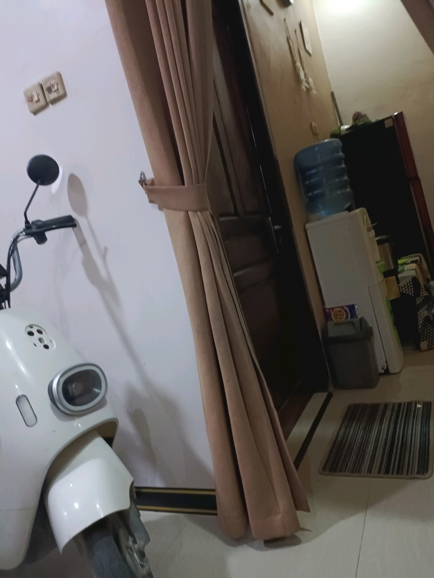 The image depicts an indoor scene, likely a hallway or entryway, with a scooter parked to the left and a water dispenser and refrigerator on the right. A tan curtain hangs prominently in the center, partially obscuring a doorway. On the wall to the left of the curtain, two light switches are visible. The flooring appears to be tiled. The overall lighting suggests an interior space with artificial light sources. The location is identified as Rancang Kencono, Indonesia. There are no people visible in the image.