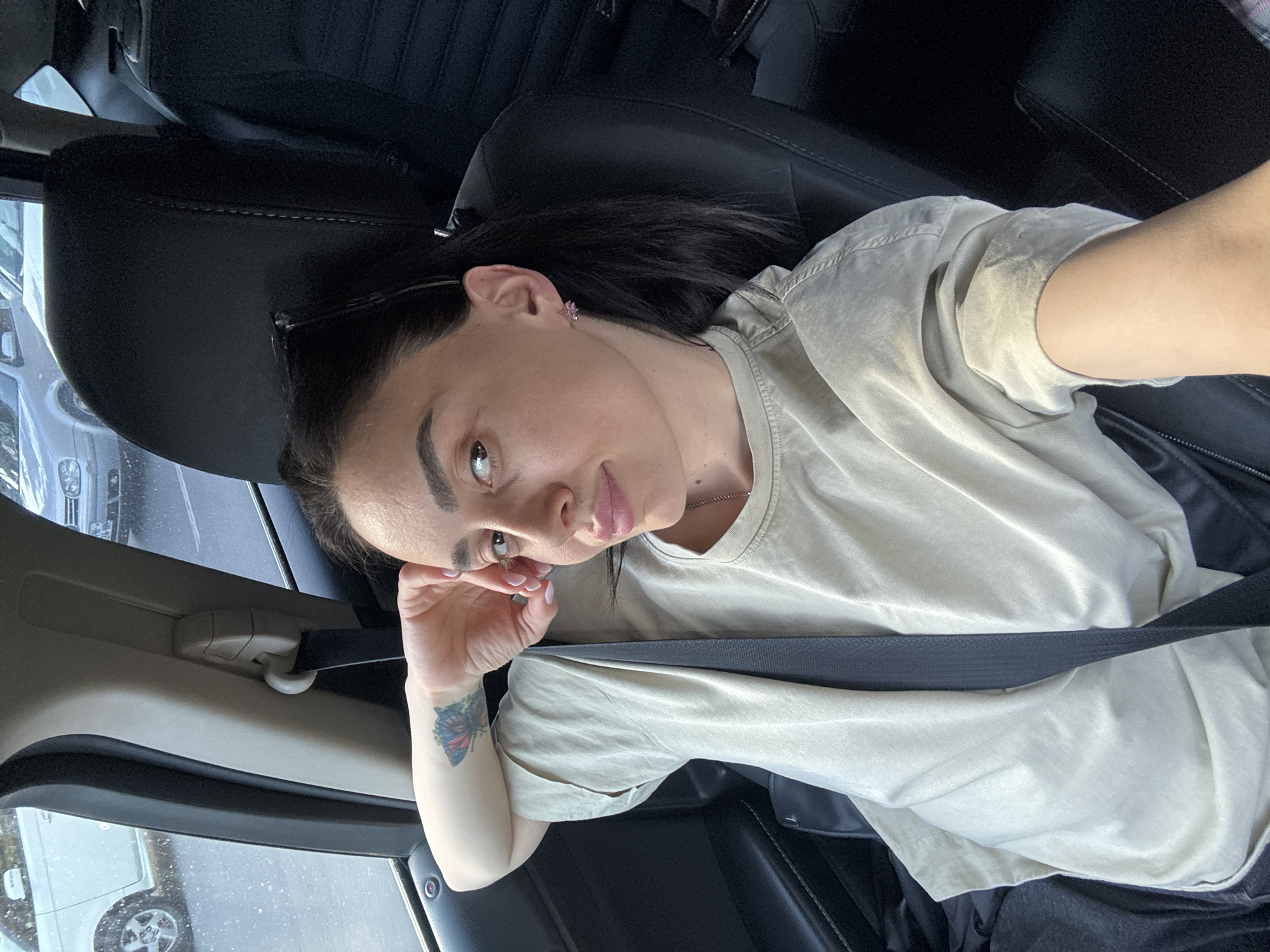 A young adult female is depicted in a medium-close up shot, positioned in the driver's seat of a car, looking directly at the camera. The image is taken at a slight upward angle, with her head tilted. She has dark hair, dark, well-defined eyebrows, and light-colored eyes. She is wearing a plain, light beige or tan short-sleeved t-shirt. Small pink stud earrings are visible, and she has a simple necklace with a small pendant. Sunglasses are pushed up onto her head.
Her left hand is gently resting near her face, revealing light-colored nail polish and a colorful butterfly tattoo on her left forearm/wrist, featuring shades of blue, red, and orange. She maintains a calm, neutral expression.
The interior of the car features dark upholstery on the seats, and a dark seatbelt crosses diagonally across her chest and lap. Through the car windows, other vehicles are visible, suggesting an urban or suburban street scene. The lighting indicates daytime, and the wet appearance of the road surface through the window suggests either recent rain or overcast conditions. The overall impression is a casual, everyday moment captured inside a vehicle. No visible text is present in the image.