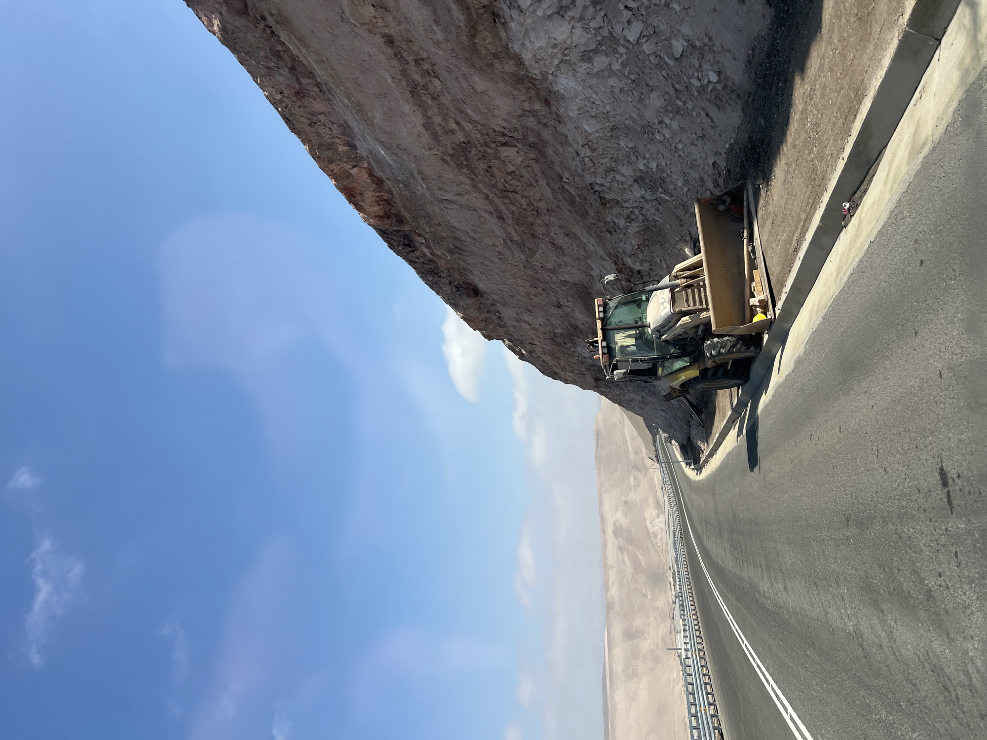 The image shows a view of a road in Arica, Chile. The road runs along the base of a cliff. A construction vehicle, possibly a loader or bulldozer, is parked on the shoulder next to the cliff. The roadway has a painted center line and a metal safety barrier on the far side. The sky is clear and blue with some clouds. Beyond the road, there is a desert landscape. A person wearing a white helmet can be seen on the curb edge, facing away. The image appears to be taken during daylight.