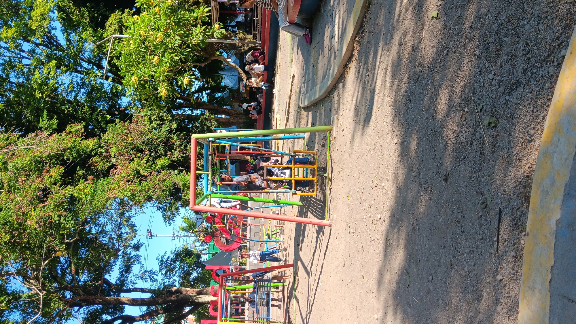 The image depicts a sunny outdoor scene at a playground in Nganjuk, Indonesia, likely a public park. A vibrant, multi-colored swing set is a central feature, with several children and at least one adult present. Some children are on the swings, while others appear to be standing around the equipment. In the background, more people are visible, some sitting on a raised concrete bench or platform, and others standing or walking. Lush green trees with some foliage and fruit, along with a clear blue sky, dominate the upper portion of the image, suggesting a warm, pleasant day. The ground of the playground is covered in gravel and dirt, with shadows cast by trees indicating the time of day. A paved pathway with a yellow and blue border is also visible in the foreground, leading into the park area.