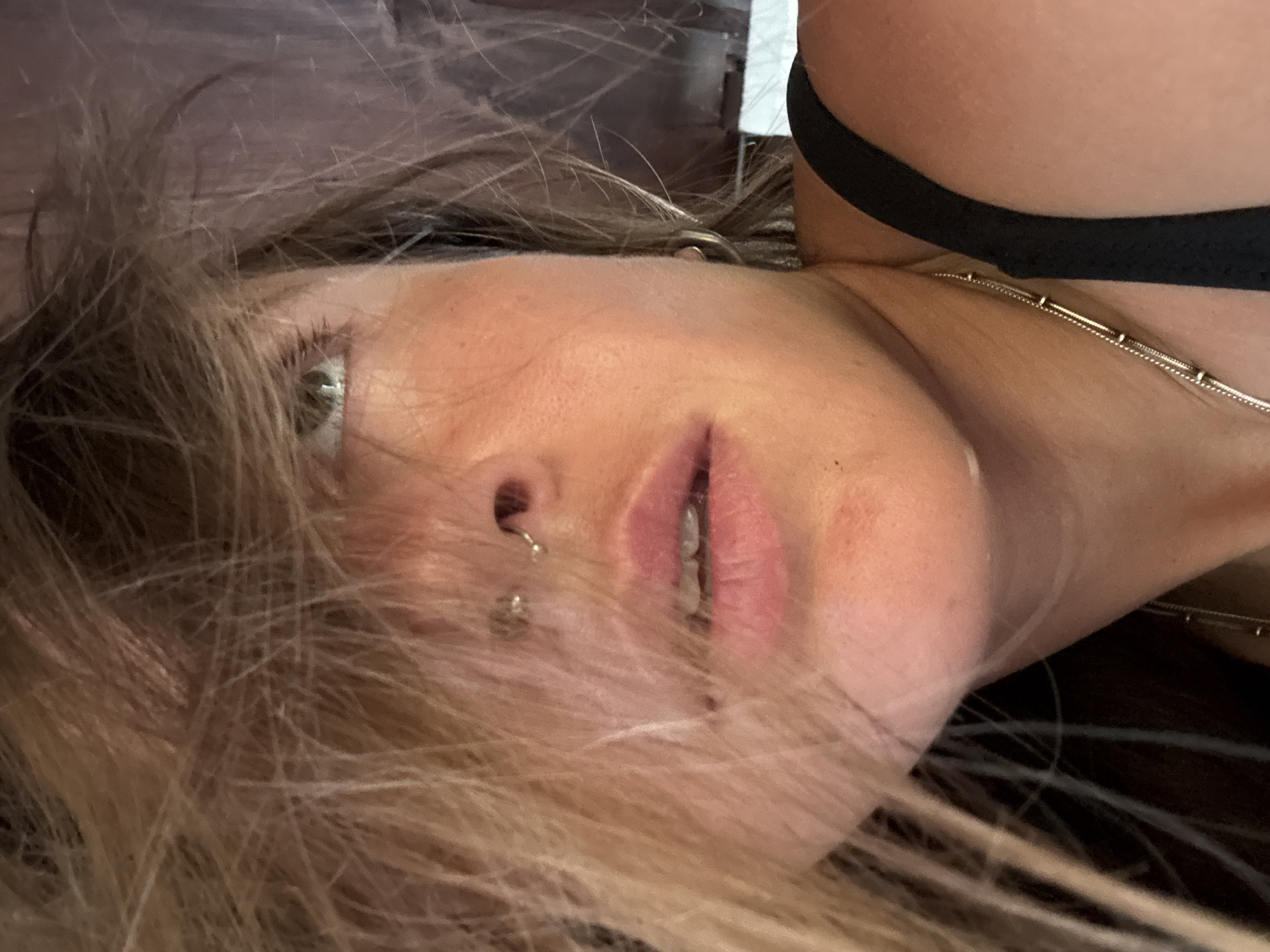 The image is a close-up, low-angle shot of a woman's face, partially obscured by her blonde hair. She is looking upwards and to her right. Her nose has a septum piercing. Her lips are slightly parted, revealing her teeth. She is wearing a black tank top and a thin silver necklace with small gold beads. The background is dark and out of focus, appearing to be a wooden surface. The lighting is warm and natural, suggesting an indoor setting during the daytime. The overall mood is casual and intimate.
