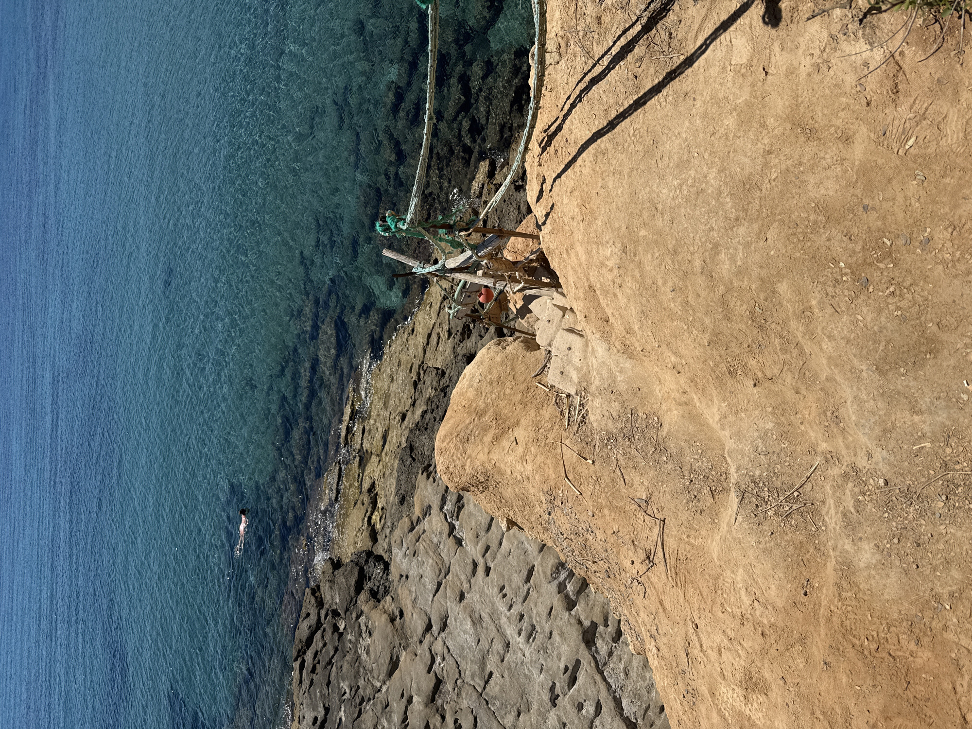 This is a vertical, aerial shot of a coastline with clear turquoise water. The scene features a rugged, sandy cliff face on the right, casting long shadows that indicate it is likely late afternoon or early morning. The water transitions from a lighter blue further from shore to a deeper, clearer turquoise closer to the rocky shore, allowing the seabed to be visible.
In the water, a single person is swimming, appearing as a small, dark silhouette. The water meets a rough, rocky shoreline made of dark grey, textured rocks. Further up the cliff, a makeshift wooden structure with ropes and a red buoy is secured to the cliff face, possibly for access or to support something. The texture of the cliff is sandy and dry, with some scattered twigs and debris. The location appears to be a natural, undeveloped coastal area. No text is visible in the image.