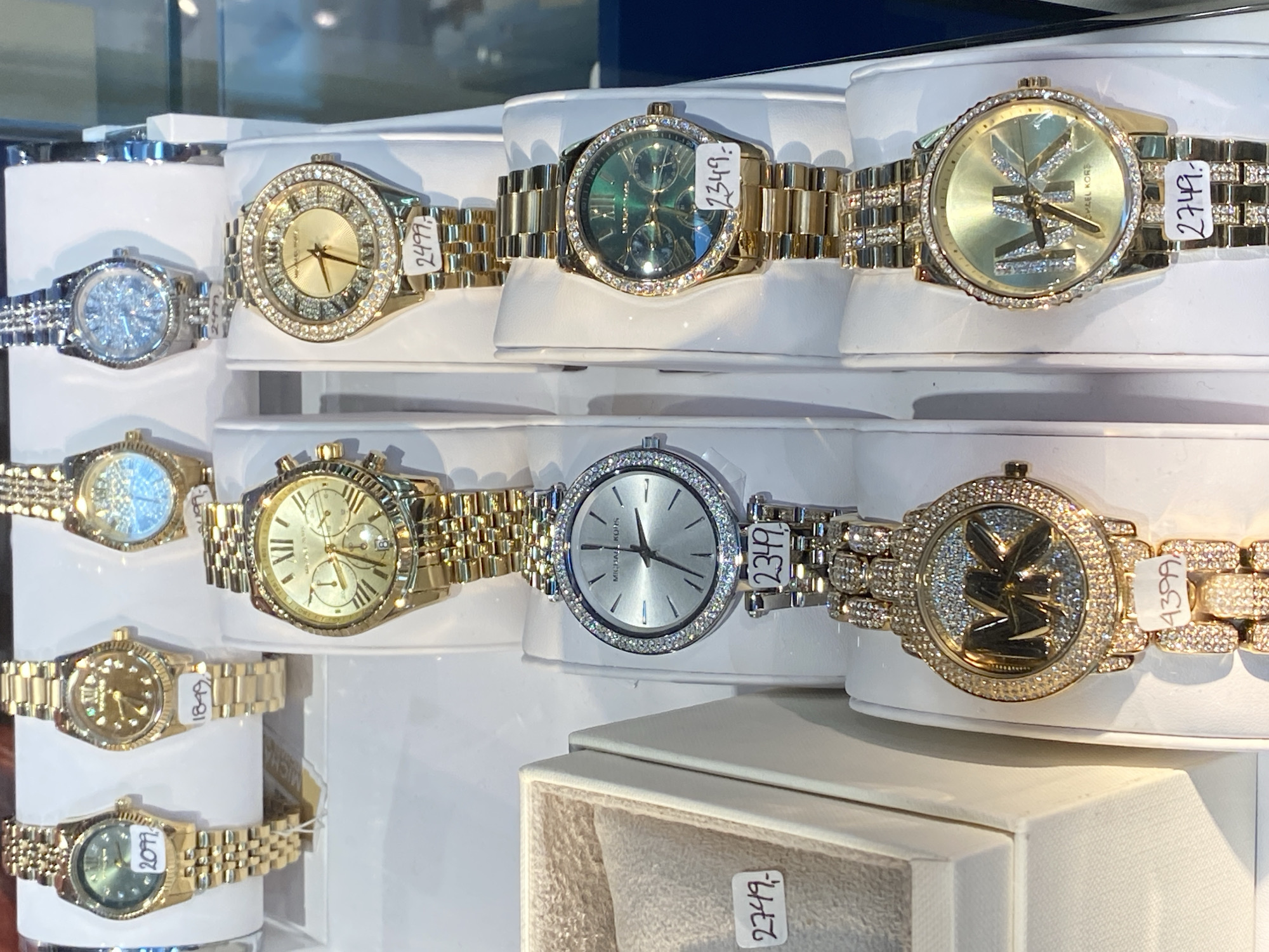 The image shows a display of Michael Kors watches in a store located in Herning, Denmark. The watches are arranged in rows on white stands within a glass display case. The watches are predominantly gold-toned with some silver-toned options. Many of them feature sparkling embellishments such as rhinestones or diamonds around the watch face and on the bands. Price tags are visible on some of the watches. The lighting suggests an indoor retail environment. No people are visible in the image.