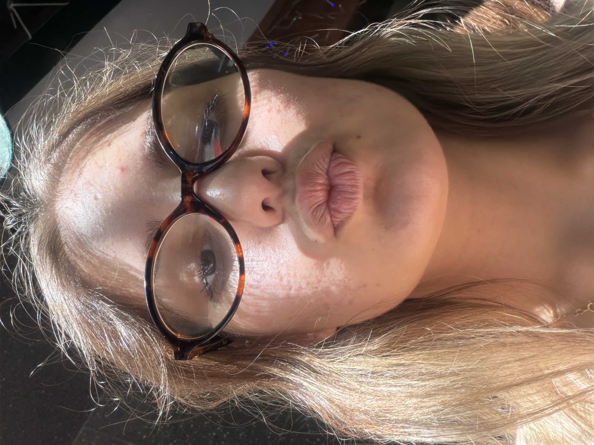 The image shows a close-up, low-angle selfie of a young Caucasian woman with blonde hair and glasses. She is making a kissing face, with her lips puckered. The lighting suggests she is outdoors, possibly near a window or in direct sunlight, as evidenced by the glare on her face and the dappled light. Her skin appears to have some freckles and minor blemishes. She is wearing tortoise-shell framed glasses and her hair is down, framing her face. A gold necklace is partially visible around her neck. The background is dark and out of focus, with some blurry blue lights visible on the right side, possibly indicating ambient lighting or decorations. No specific location in Burgas, Bulgaria can be identified from the visual information in the image.