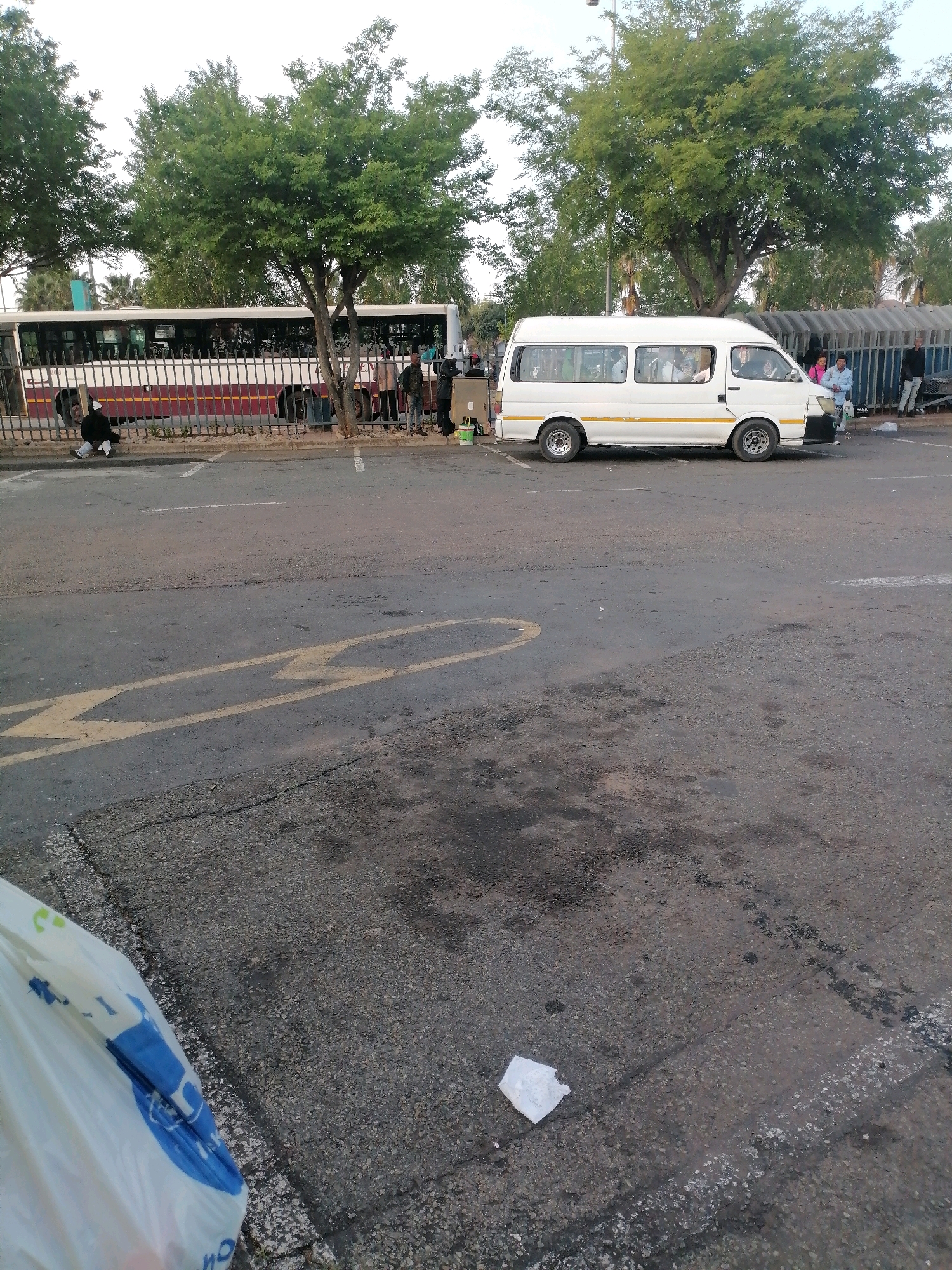 In Emfuleni Local Municipality, South Africa, a white minibus taxi is parked in a lot next to a bus. A person is seated on the ground in front of the bus, facing away from the camera. Trees line the perimeter of the lot, and in the background, several people are visible near the bus. The ground is paved, with visible road markings, and there is some litter present. The lighting suggests it is daytime.