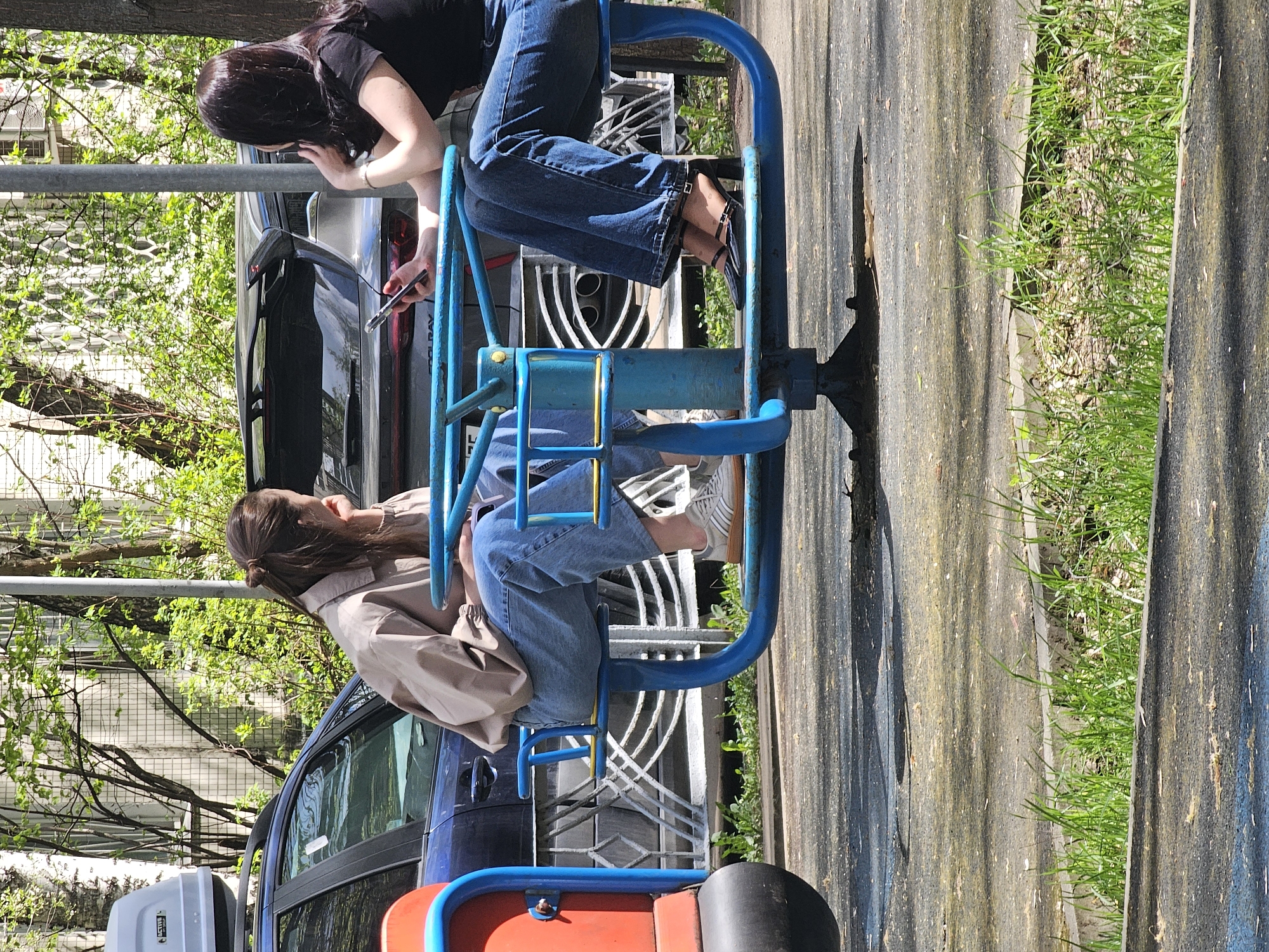 Two women are on a blue, red, and black playground roundabout in Khimki, Russia. One wears a black shirt and jeans and holds a phone near a black car. The other has on a tan jacket and jeans. The roundabout is on an asphalt surface with a strip of grass and concrete panels alongside it. There are cars parked nearby and trees with green leaves in the background.