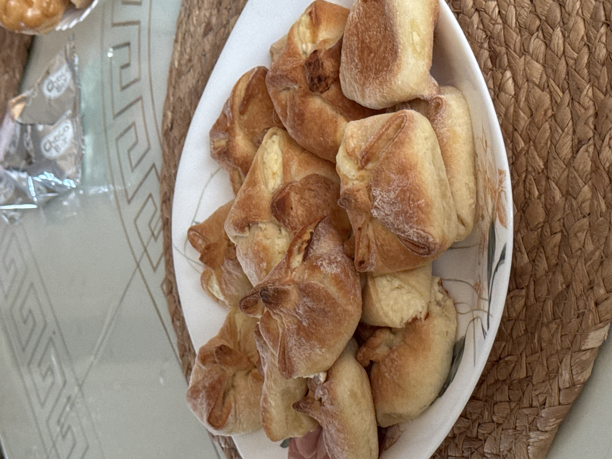 The image presents a close-up, high-angle view of a white oval dish filled with baked goods. The baked items appear to be golden-brown and irregularly shaped, possibly filled pastries or small buns. The dish sits on a textured, woven placemat of brown color. In the background, a portion of a table is visible, with a metallic surface and a geometric design, as well as a wrapper for "Choco" candies. The location, Izhevsk, Russia, is provided in the prompt.
