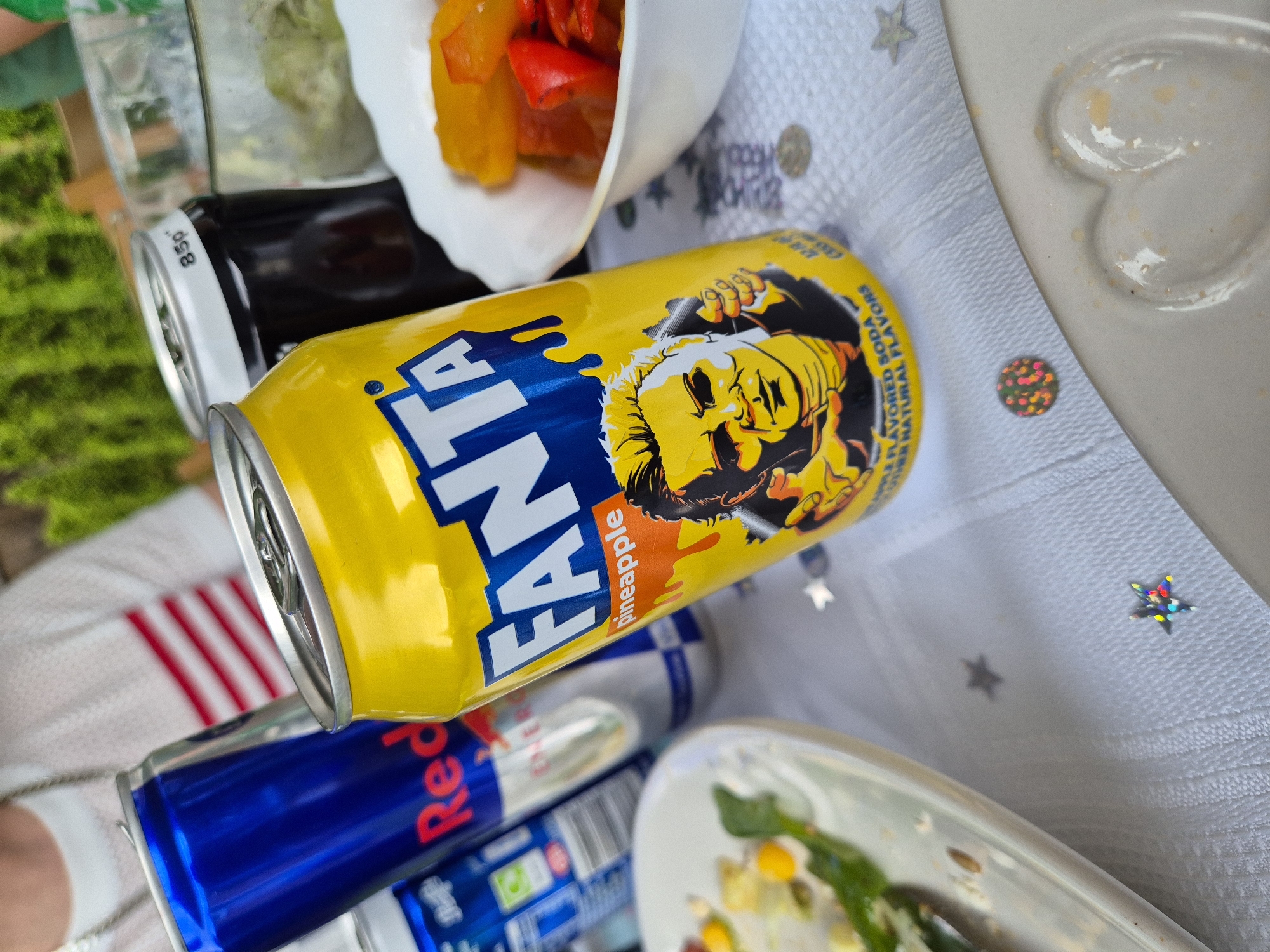 The image shows a table setting outdoors. There are cans of Fanta Pineapple, Coca-Cola, and Red Bull. A bowl of cooked vegetables is visible. The setting appears to be a casual meal or picnic. The location, West Suffolk, United Kingdom, is inferred from the packaging of some items possibly available locally.