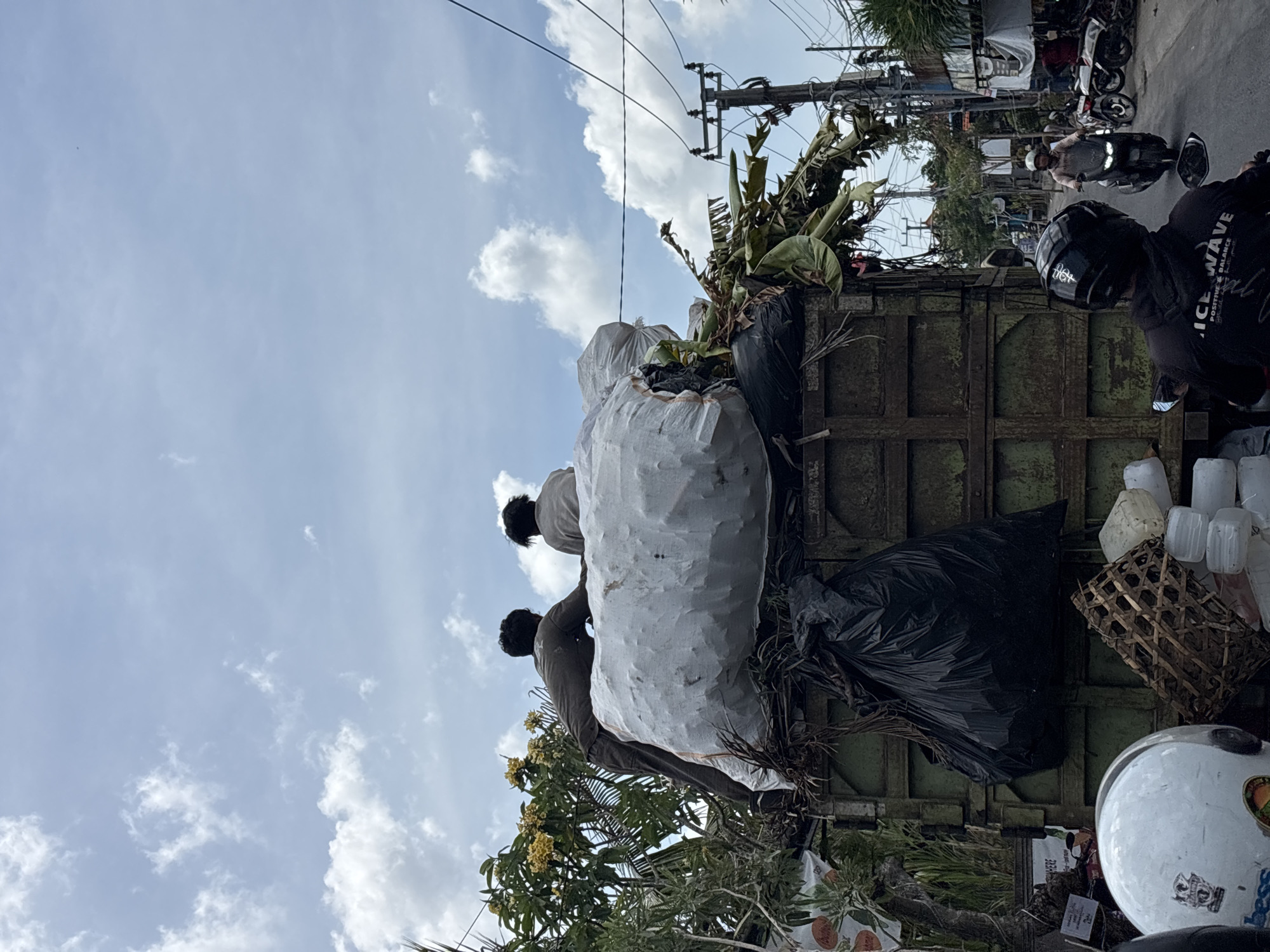 The image shows the back of a green, rusted truck filled with refuse, including large white sacks, black plastic bags, banana leaves, and assorted plastic items. Two men are standing on the truck, handling the refuse. In the background, there are utility poles, a partly cloudy sky, and other motorbikes on the road. The scene is located in Mengwi, Indonesia.