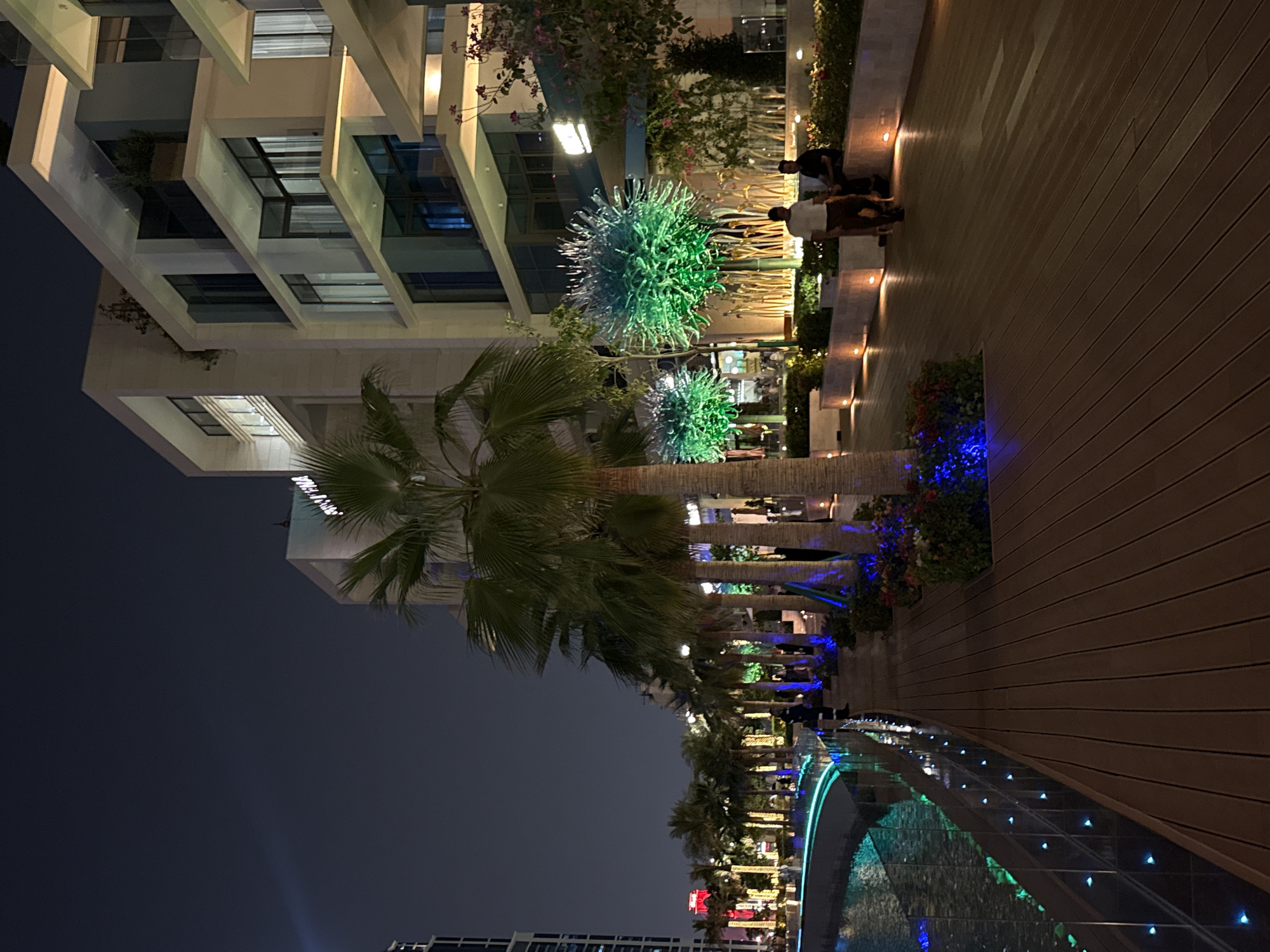 The image shows a nighttime scene in Doha, Qatar. A modern, multi-story building is visible on the left, featuring cantilevered balconies and large windows. In the foreground, a row of palm trees lines a walkway, with ornamental lighting illuminating the area. The walkway is paved with wooden planks and bordered by garden beds. A water feature with integrated lights is present along the edge of the walkway. Some people are visible near the building on the right side of the frame. In the distance, other buildings and lights indicate an urban environment.