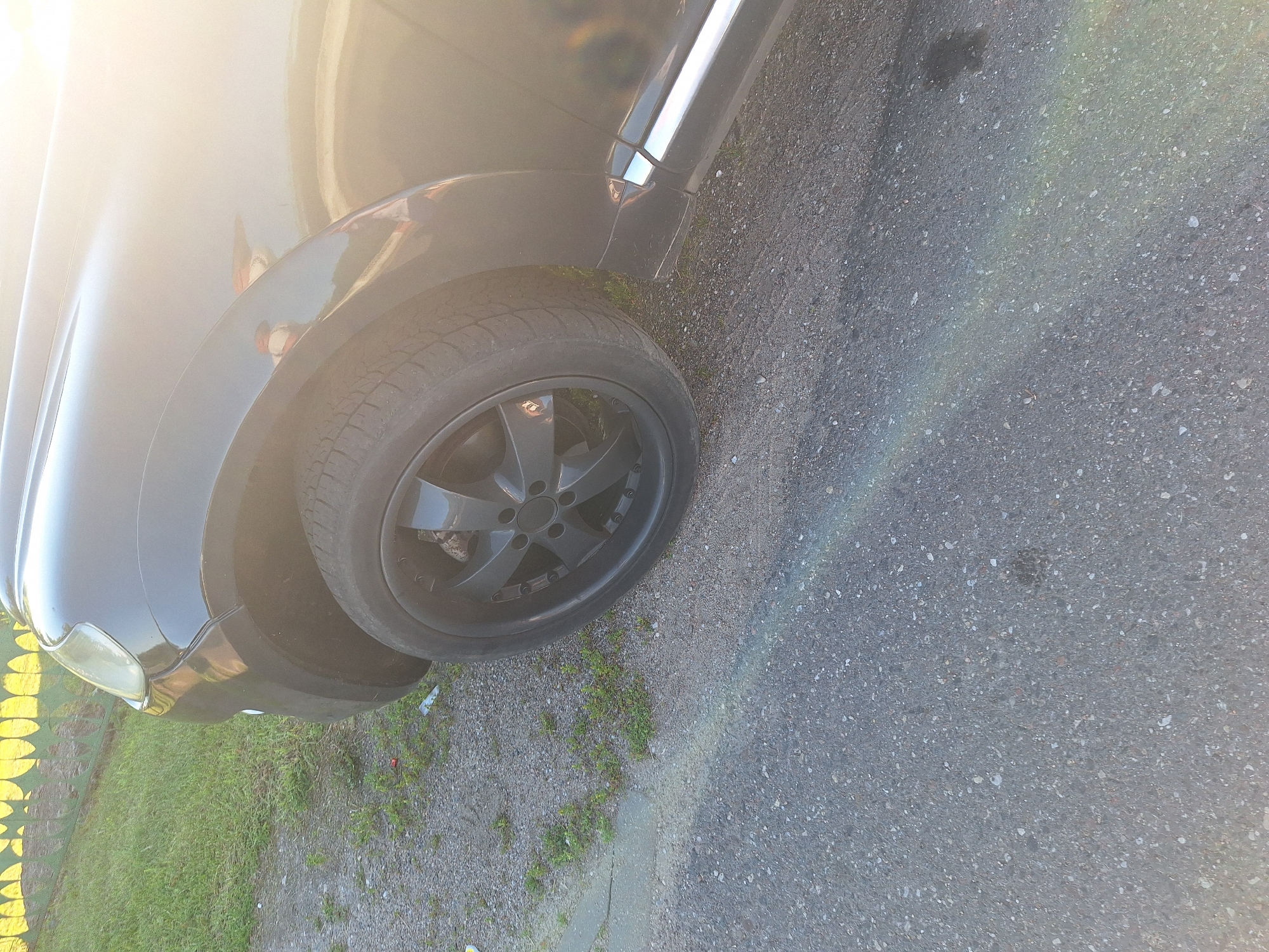 This is a close-up, eye-level, outdoor photograph of the front passenger side wheel and fender of a black SUV. The vehicle is parked on an asphalt surface with a gravel and grass verge to the left. A green metal fence with yellow circular designs is visible in the upper left quadrant, bordering a grassy area. The setting appears to be a residential area in Цімкавіцкі сельскі Савет, Belarus, based on the context. The sun is low and to the right, creating a strong glare and lens flare across the vehicle's body and a faint rainbow effect on the asphalt. The tire is black with a multi-spoke dark grey rim. The fender is black and curves over the wheel well. The vehicle's headlight and part of the hood are visible. There are no people or other distinguishing features in the image.