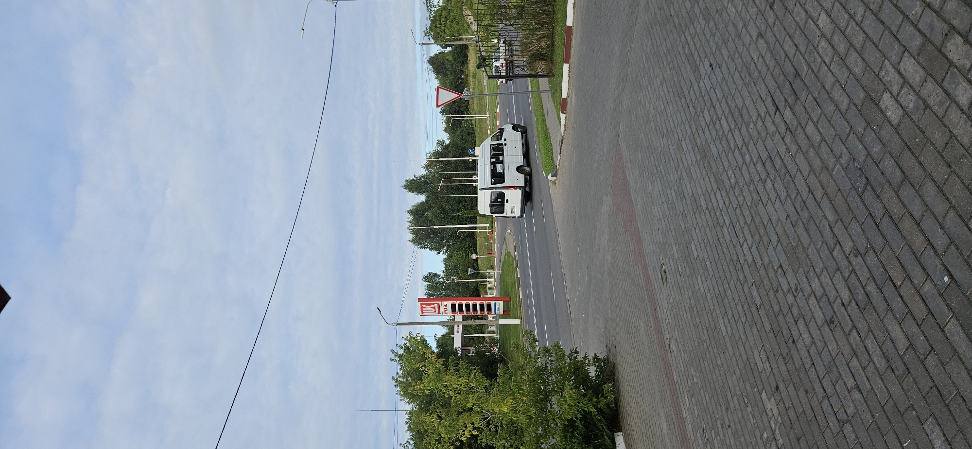 The image shows a view of a street intersection in Vitsebsk, Belarus, featuring a white minibus driving on the road. A gas station with a prominent red and white Lukoil sign is visible on the left side of the image. Trees and streetlights are scattered throughout the scene. The ground in the foreground is paved with grey cobblestones. The sky is cloudy.