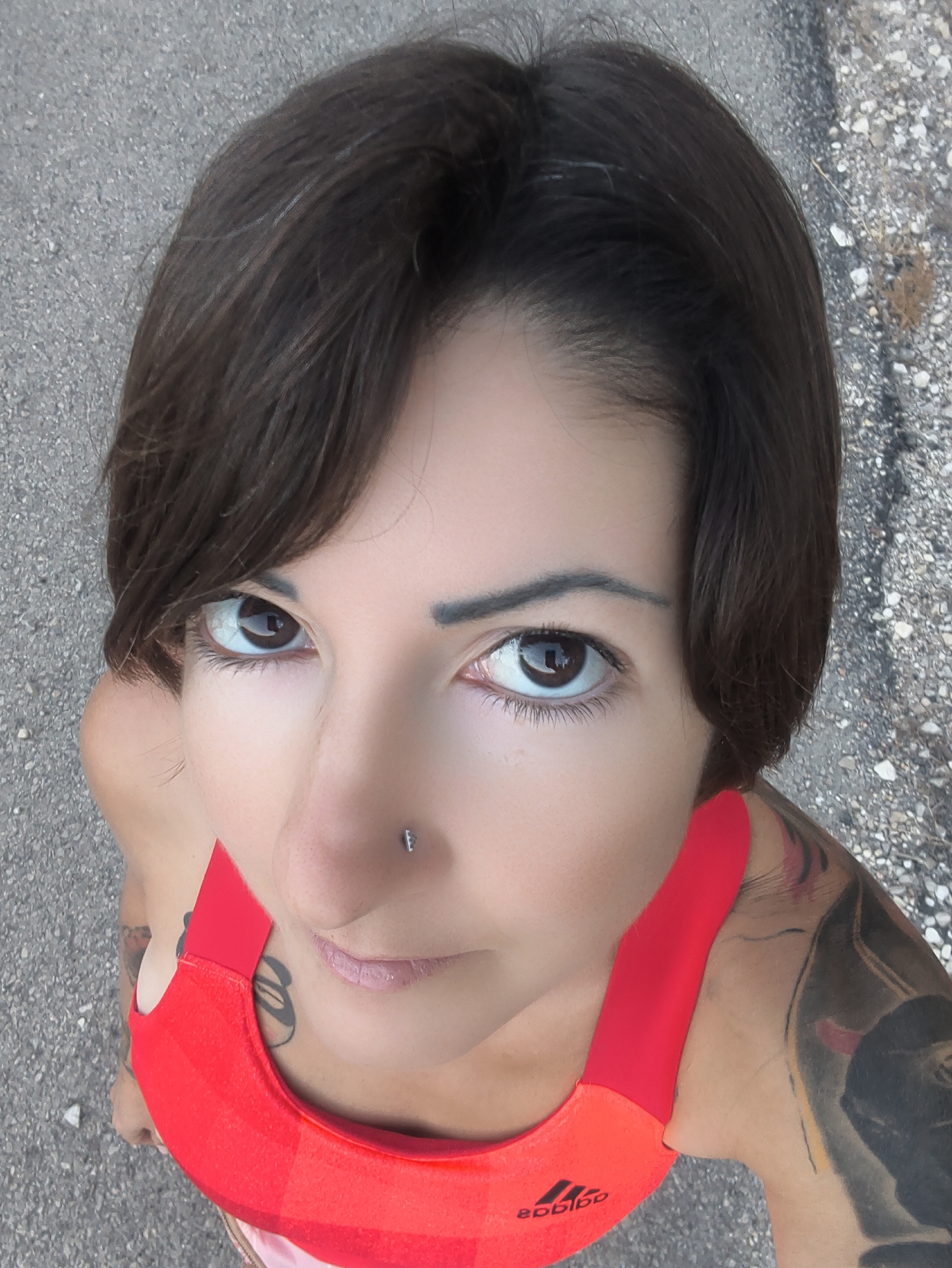 A woman with short, dark brown hair and brown eyes is captured in a low-angle shot. She is wearing a red sports bra with the Adidas logo visible on it. She has a nose ring and tattoos on her arms and chest. The background is a textured, asphalt-like surface with small stones. The image appears to have been taken outdoors. The location is specified as the Lachish Regional Council, Israel.