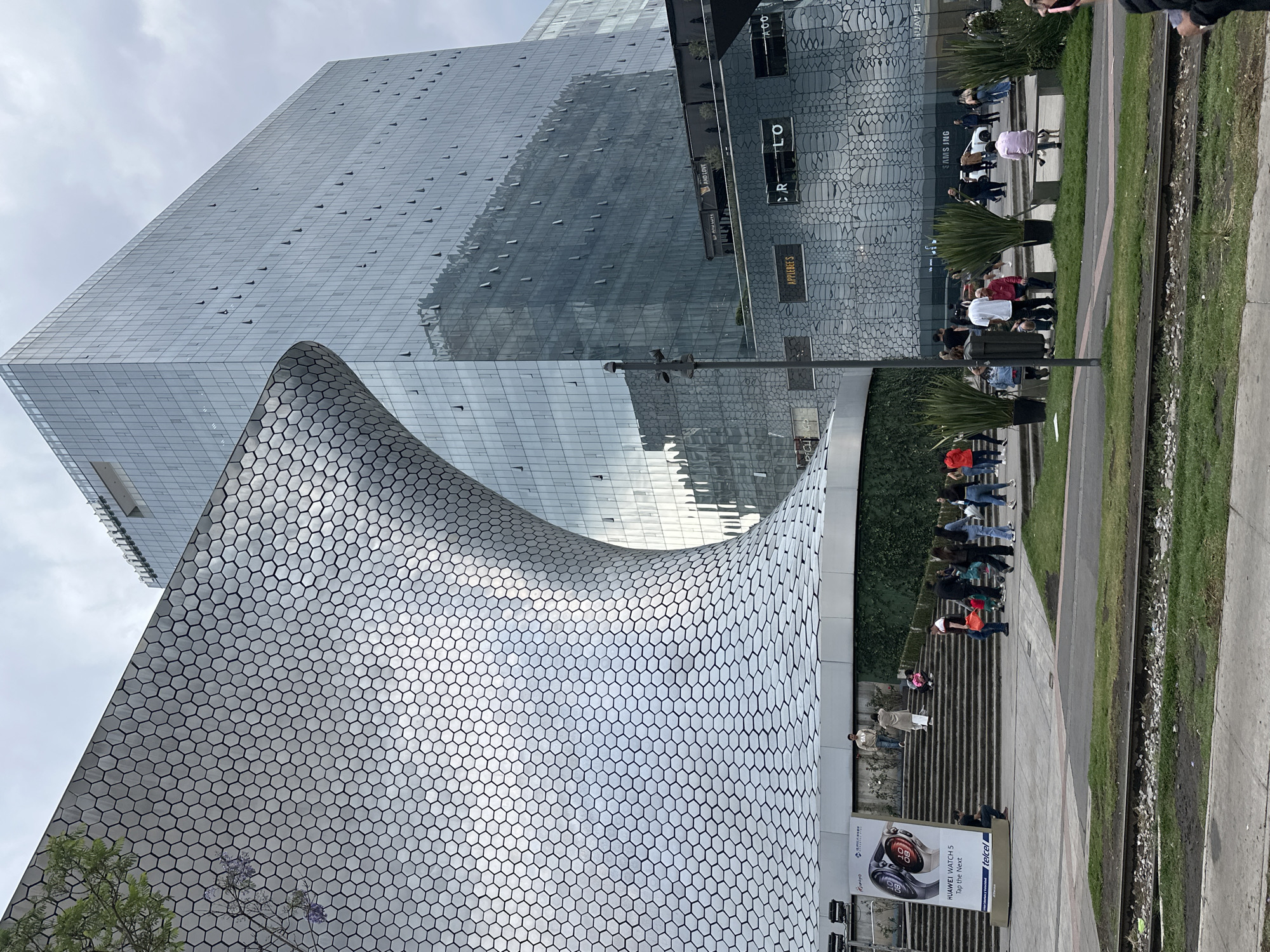 The image captures a street scene in Mexico City, Mexico, showcasing a modern architectural landscape. The Soumaya Museum is prominently featured, with its unique, curvilinear structure covered in hexagonal tiles. Adjacent to the museum stands a tall, reflective glass building, possibly an office or residential tower.
A line of people is visible along a raised walkway beside a narrow strip of green grass. The walkway appears to be elevated, with stairs leading down to a lower level. Along the base of the Soumaya Museum, signage and advertisements are displayed, including a large ad for a Huawei watch with Telcel branding. Commercial establishments such as Applebee's and other retail stores are also visible on the side of the glass building.
The scene is set on an overcast day, casting a soft light on the buildings and surrounding environment. The image offers a glimpse into the contemporary urban landscape of Mexico City.