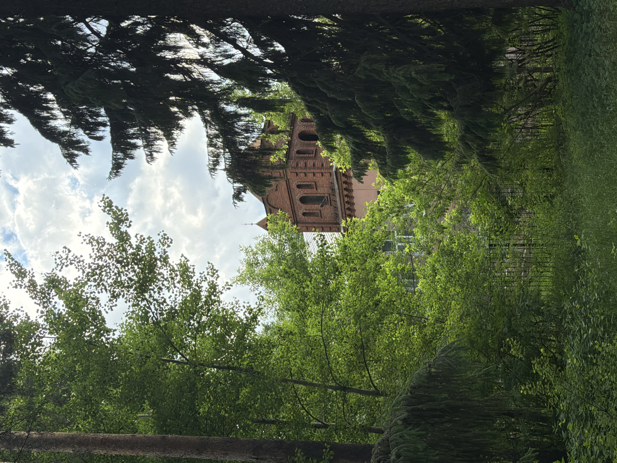 The image shows a brick building in Votkinsk, Russia, partially obscured by trees. The building features a tower with arched windows. The sky is visible through gaps in the trees, showing clouds. The surrounding area appears to be a park or wooded area.