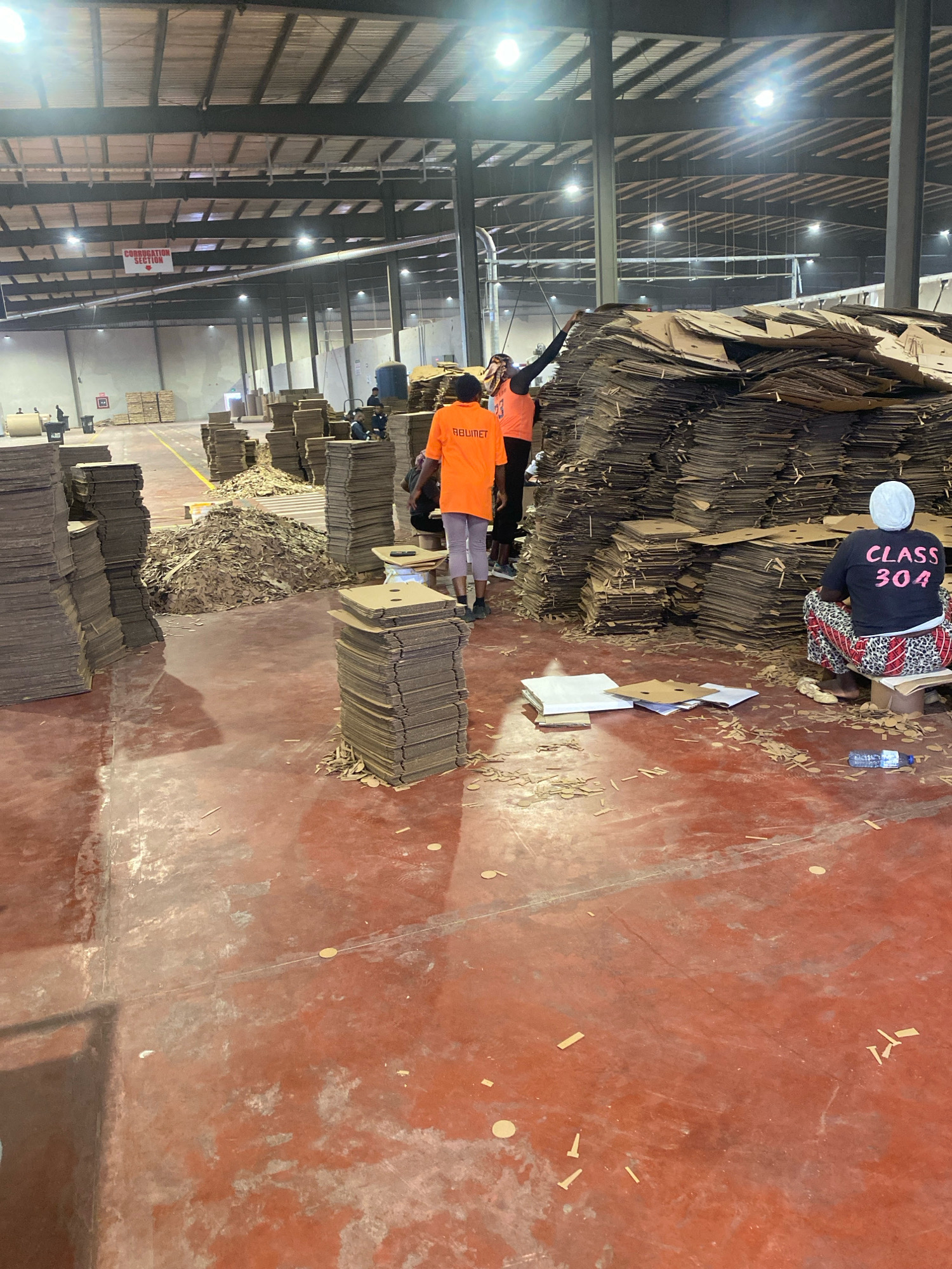 This is an indoor photograph taken at a cardboard manufacturing or recycling facility in Abuja, Nigeria. The large industrial space has a high ceiling with exposed beams and lighting fixtures. The floor is a reddish-orange concrete.
In the foreground and midground, stacks of corrugated cardboard are piled high, some neatly arranged and others in disarray. Small pieces of cut cardboard are scattered across the floor.
Three individuals are visible. In the right midground, a person wearing a white headscarf and a black t-shirt with "CLASS 304" printed on it in pink is seated on a cardboard box. In the left midground, two people are visible. One is wearing an orange t-shirt with "REBINET" printed on it and the number "23" below. This person is standing and appears to be engaged in work. Another person is partially visible behind them, also wearing an orange t-shirt.
In the background, more stacks of cardboard are present, and other individuals can be seen working in the distance. A red sign hangs from the ceiling in the far background, reading "CORRUGATION SECTION" in white lettering with an arrow pointing to the right. The overall scene depicts an active workspace involved in the processing of cardboard.