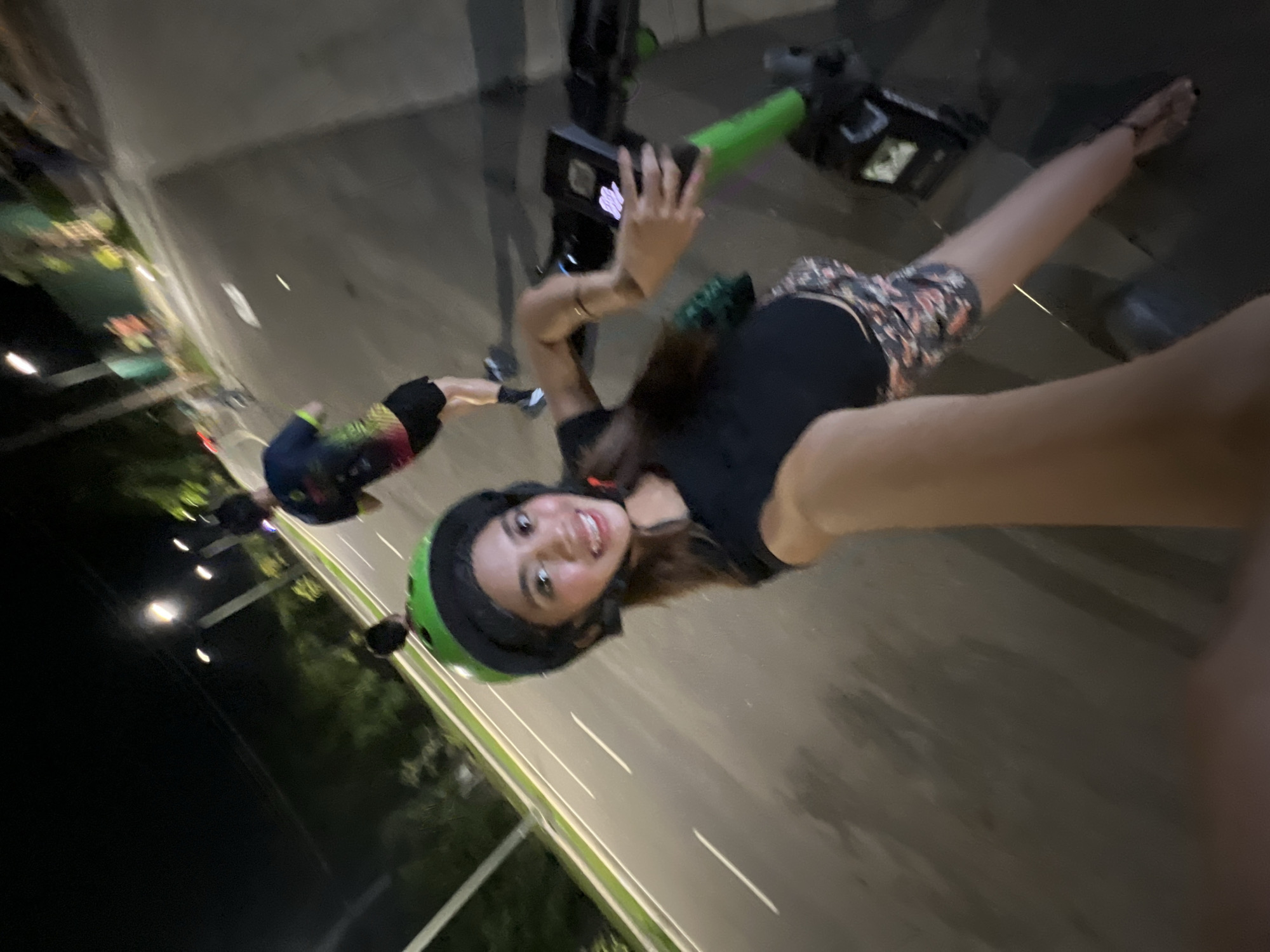 The image is a selfie taken by a young woman at night in Davao City, Philippines. The woman is smiling broadly, looking directly at the camera, and holding the phone in her outstretched right hand. She has long, dark hair and is wearing a black top, floral-patterned shorts, and a green and black helmet. Her left arm, adorned with a bracelet, is also visible.
She appears to be on an electric scooter, with its green handlebars and part of the base visible to her right. Another person, also likely on an electric scooter and wearing a helmet, is partially visible in the blurred background to the left. The scene is outdoors, on a paved surface that resembles a sidewalk or a dedicated lane next to a road, indicated by white markings. Streetlights illuminate the area, and trees and greenery are visible in the distance. The time of day is clearly night. The overall impression is one of leisure and enjoyment, possibly during an evening ride. No discernible text is visible in the image.