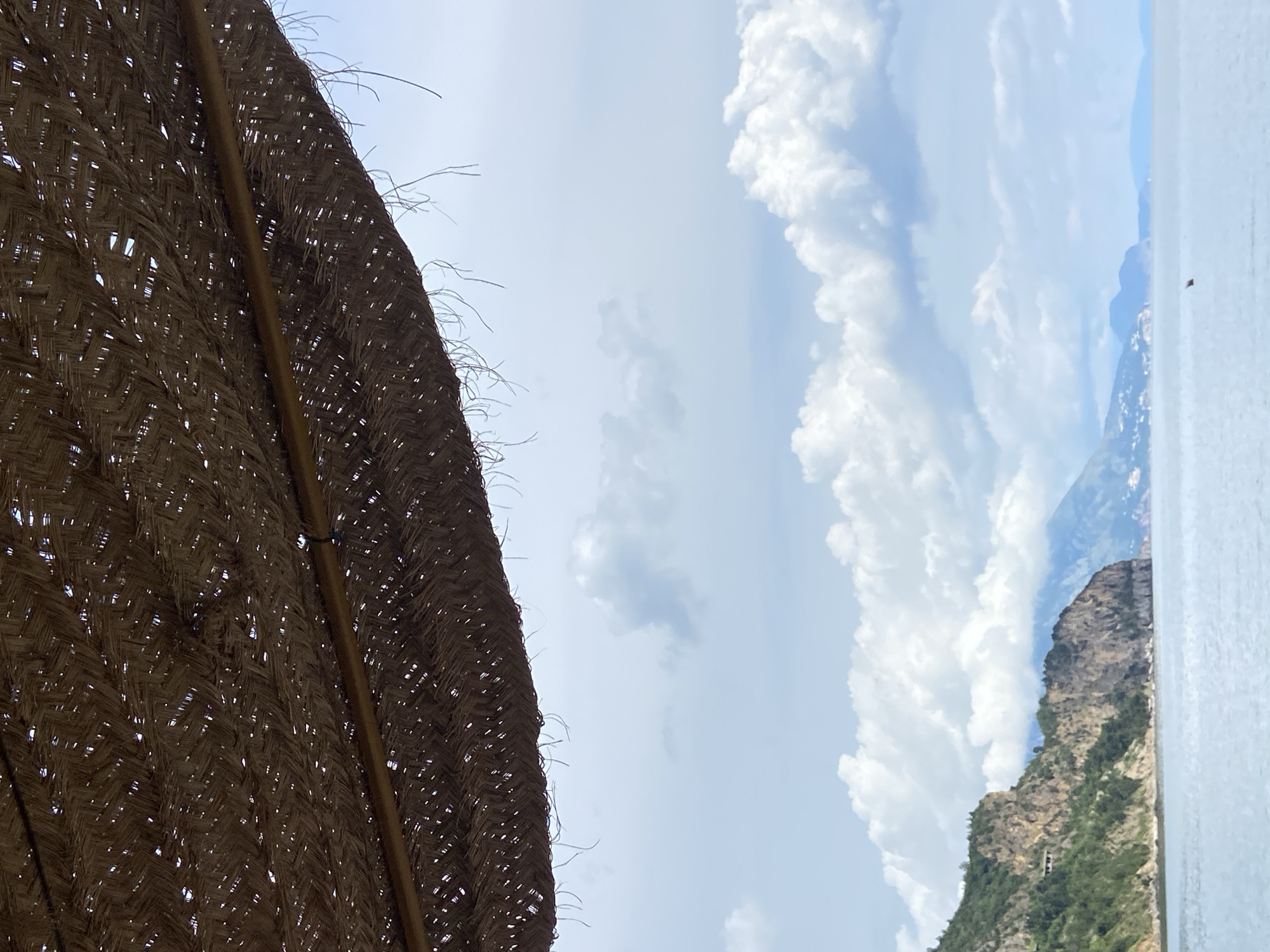 The image shows a view from under a straw beach umbrella. The sky is visible, with puffy white clouds scattered across a blue backdrop. To the right, a portion of a rocky mountain covered in green vegetation is seen. A sliver of what appears to be the sea or a large body of water is also visible on the far right. The scene suggests a coastal location in Montenegro, possibly near Prijevor, but without further context, a definitive identification of the precise location is difficult. The image features no people and depicts a static landscape scene.