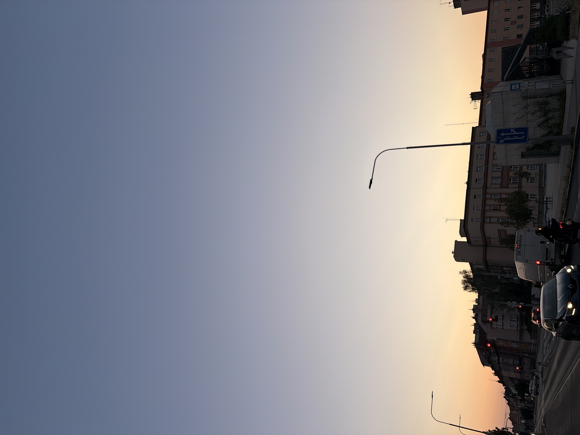 The image depicts a street scene in Pilsen, Czechia, during dusk. The sky transitions from a deep blue at the top to a pale orange near the horizon, indicating the setting sun. A street light with a curved arm is prominently visible in the middle ground on the right.
On the left side of the image, several vehicles are on the road, including a dark-colored car in the foreground, a white van further back, and a motorcycle with a rider. Traffic lights are visible, some showing red. Buildings line the street, featuring multiple stories and windows. A signpost with blue directional arrows is positioned near the street light. The overall environment appears to be an urban setting with ongoing traffic.