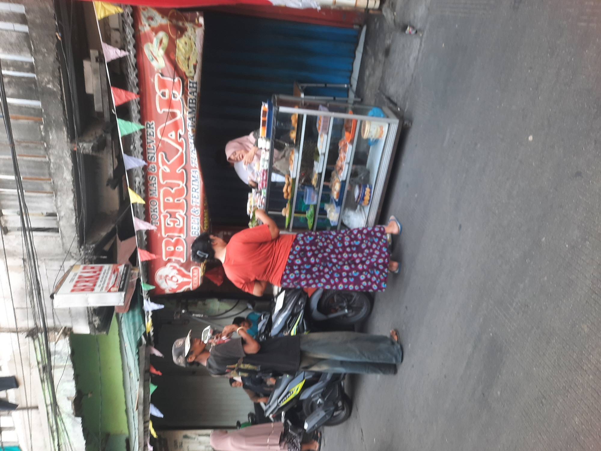 This image depicts an outdoor scene in North Jakarta, Indonesia, likely in a commercial or residential street setting. In the foreground, a man wearing a black t-shirt, a cap, and loose-fitting pants stands next to a motorcycle. He appears to be holding a phone or a similar device. To his right, a woman in a red t-shirt and a patterned skirt is standing in front of a glass display case filled with various food items. Behind the food stall, a woman in a headscarf is visible, seemingly attending to the stall. The stall has a red awning with text that reads "BERKAH" in large letters, suggesting it is a business. Colorful triangular flags are strung above the awning. The background shows a building with a concrete facade and an open doorway covered by blue curtains. The ground is paved asphalt. The overall atmosphere suggests a bustling street-level commercial activity in a local neighborhood.