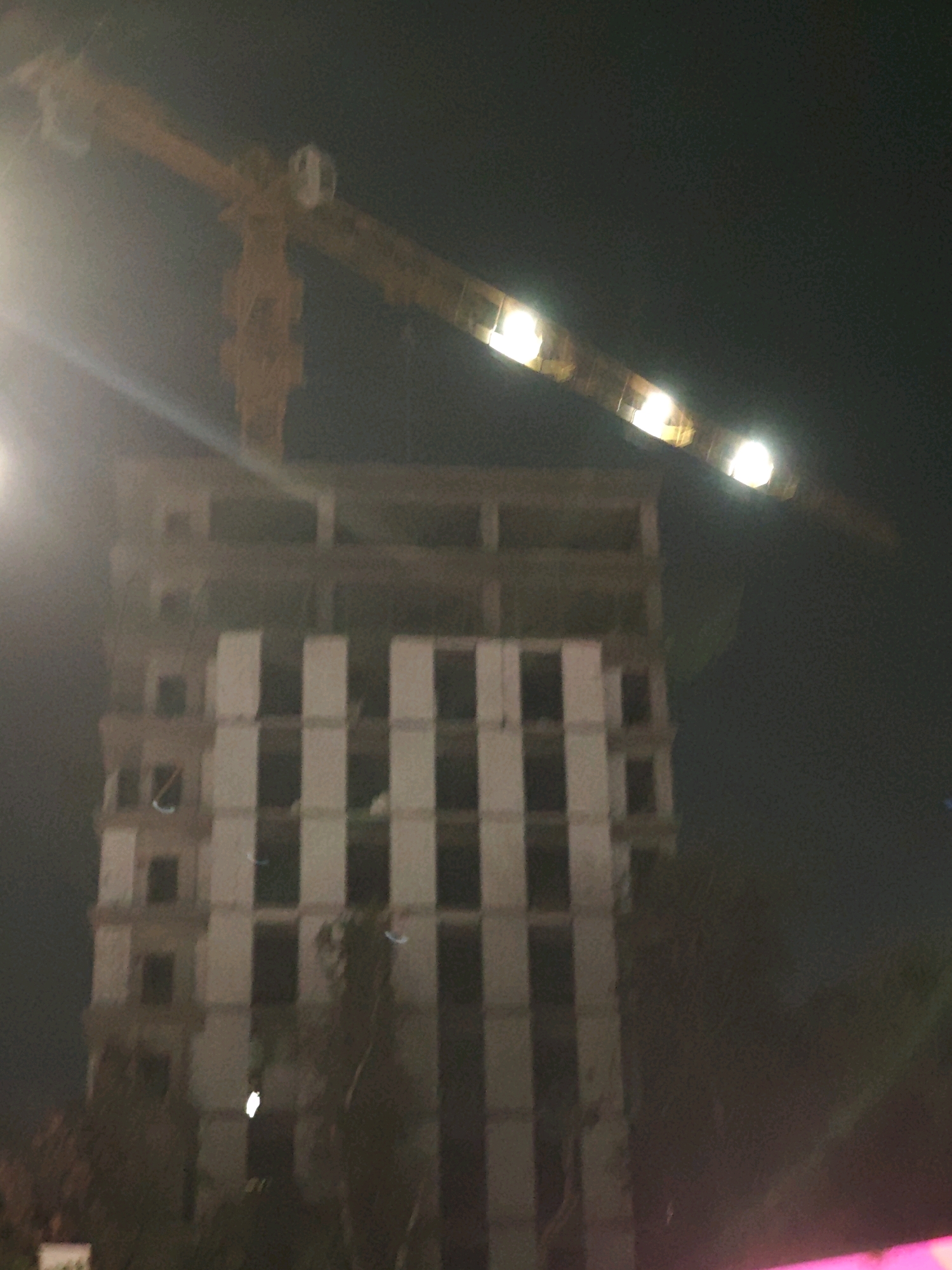 The image shows a night scene of a construction site in Tashkent, Uzbekistan. A large yellow tower crane is positioned above an unfinished multi-story building. The building appears to be made of concrete and has a repetitive facade with dark rectangular openings. Several bright lights illuminate parts of the crane and the upper sections of the building. The sky is dark, and some light streaks, possibly from streetlights or reflections, are visible. The lower part of the image is obscured by shadows and foliage, with hints of pink and magenta light possibly from nearby signage or event lighting.