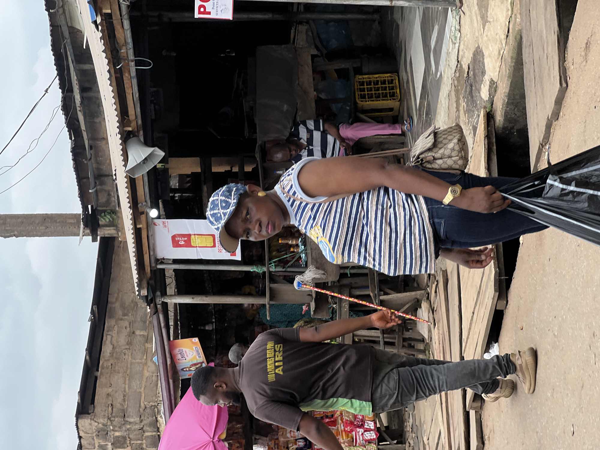 In Nkpor, Nigeria, a bustling marketplace scene unfolds under an overcast sky. The foreground is dominated by a woman in a blue and white striped sleeveless top and dark denim jeans, her stance slightly turned, holding a black plastic bag. She wears a blue and white checkered cap and a gold watch on her left wrist. To her left, a man in a brown t-shirt with yellow lettering and dark grey jeans walks away from the camera, his right hand holding a mop with a red and white handle. Behind them, other individuals are visible within the market stalls. One stall displays an Airtel advertisement, and a white loudspeaker is mounted above another. Wooden planks form makeshift pathways over a drainage channel. The overall environment suggests a commercial area with temporary structures and a lively atmosphere.