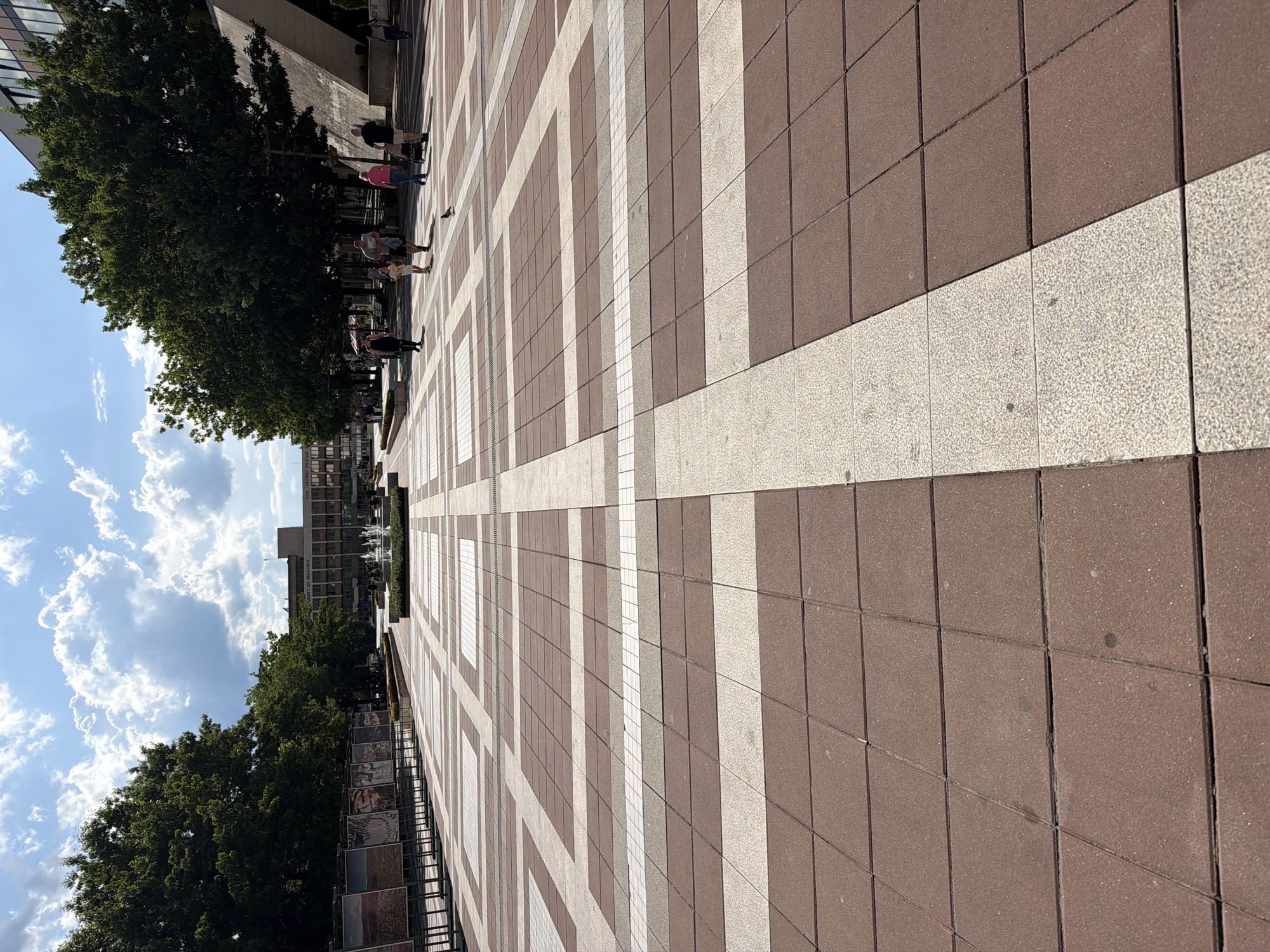 The image captures a wide-angle view of a sunlit outdoor pedestrian plaza, characterized by a striking geometric pattern on its paved surface. The ground is covered with square tiles, arranged in alternating blocks of light beige and reddish-brown.
Lush green trees line both the left and right sides of the plaza, casting distinct shadows on the pavement, indicating bright daytime sunlight. Several individuals are scattered across the area, walking or standing casually. On the left, near the trees, a small group of people in everyday attire, including one person wearing a pink top and another a black top, are visible.
In the background, modern buildings rise above the treeline. One building prominently features multiple windows, and faint text on its facade partially reads "UNIVERSITY OF," suggesting a university setting. To the right in the background, a water fountain with active jets is visible, surrounded by greenery. Along the right side of the plaza, a series of rectangular display panels are lined up, possibly an outdoor art installation or an informational exhibition.
The sky is a clear blue with scattered large, white cumulus clouds, signifying pleasant weather. The scene depicts a calm, ordinary day in a public space, likely a university campus or city square in Blagoevgrad, Bulgaria.
