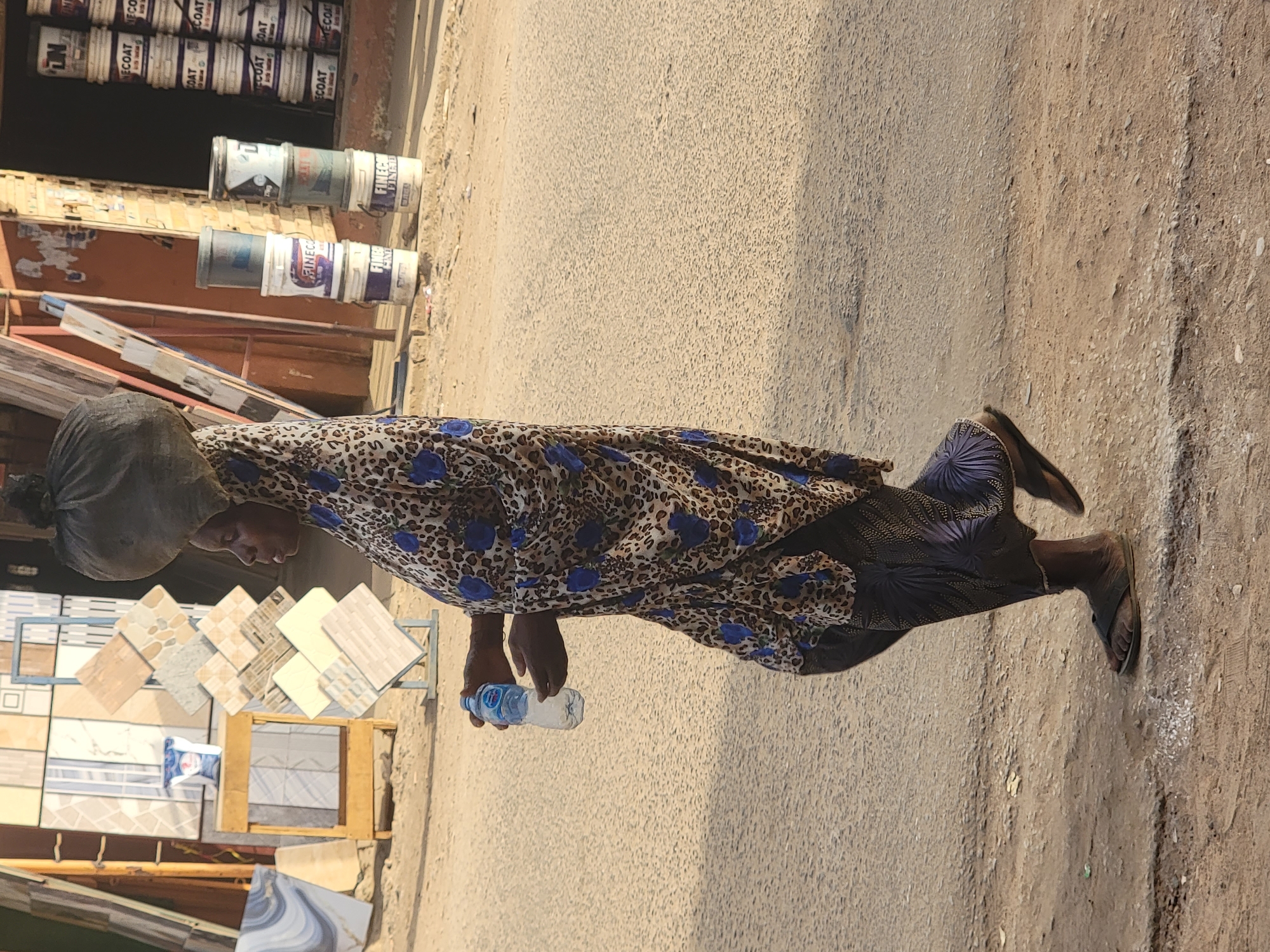 An African woman walks on a paved street next to an outdoor tile vendor and a paint vendor in Maiburiji, Nigeria. She carries something on her head and a bottle of water in her right hand. She wears a leopard-print garment with dark blue floral designs.