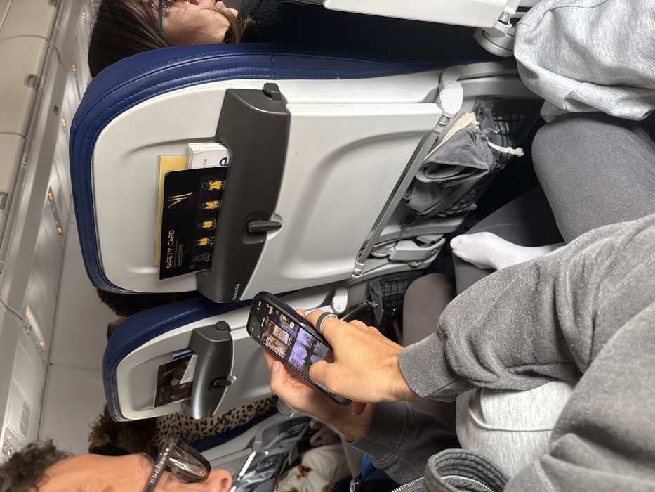 The scene is set inside an aircraft cabin, consistent with either being on the ground at or in flight to/from Lyon-Saint Exupéry Airport (LYS) in Colombier-Saugnieu, France. In the foreground, a person, discernibly male, is seated, wearing a grey hooded sweatshirt, grey sweatpants, and black-framed Gucci glasses. This individual holds a black smartphone horizontally with both hands, actively interacting with the screen, which displays multiple thumbnail images. A black and silver ring is present on the right ring finger, and the right foot, clad in a white sock, is extended. Directly in front of this person is the back of an aircraft economy seat, featuring a white plastic shell with blue trim. Its seat-back pocket contains a black "SAFETY CARD" with gold lettering and graphics of four figures in life vests, alongside a white booklet. Beneath this pocket, a mesh net holds a grey fabric item with a drawstring. To the right, another passenger is partially visible, seen in profile wearing glasses and dark attire. A third passenger, wearing a leopard-print top, is partially visible in the background to the left. The cabin overhead lighting is illuminated.