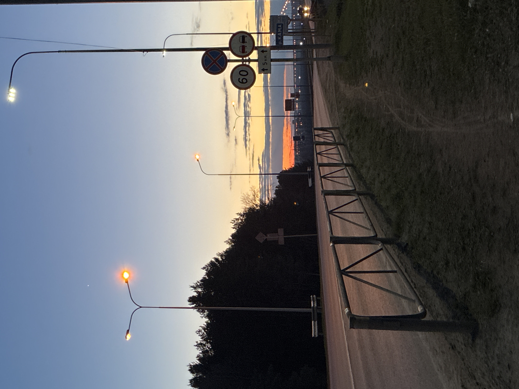The image captures a road scene at either dusk or dawn, characterized by a striking sky displaying a gradient from light blue at the top to warm orange and yellow hues near the horizon, indicative of the golden hour. The road stretches from the foreground into the distance, bordered on the right by a low metal barrier or fence alongside a grassy verge.
Several streetlights are illuminated, some emitting a bright white LED glow, while others cast a softer, yellowish light. In the mid-ground, a dense line of dark trees and foliage forms a silhouette against the colorful sky, obscuring the view of the immediate landscape beyond.
A prominent cluster of traffic signs is visible above the road. These include a "No Stopping" sign (blue circle with a red diagonal cross), a "No Overtaking for Trucks" sign (red circle with two trucks), and a circular "60" sign, indicating a speed limit. Another rectangular sign features an upward arrow and "5 km", likely denoting a distance. Crucially, a larger blue sign with white Cyrillic text clearly reads "Р. КАМА" (R. KAMA), confirming the presence of the Kama River, which aligns with the provided location context of Chaikovsky, Russia. The scene conveys a serene and quiet atmosphere as daylight fades or begins.