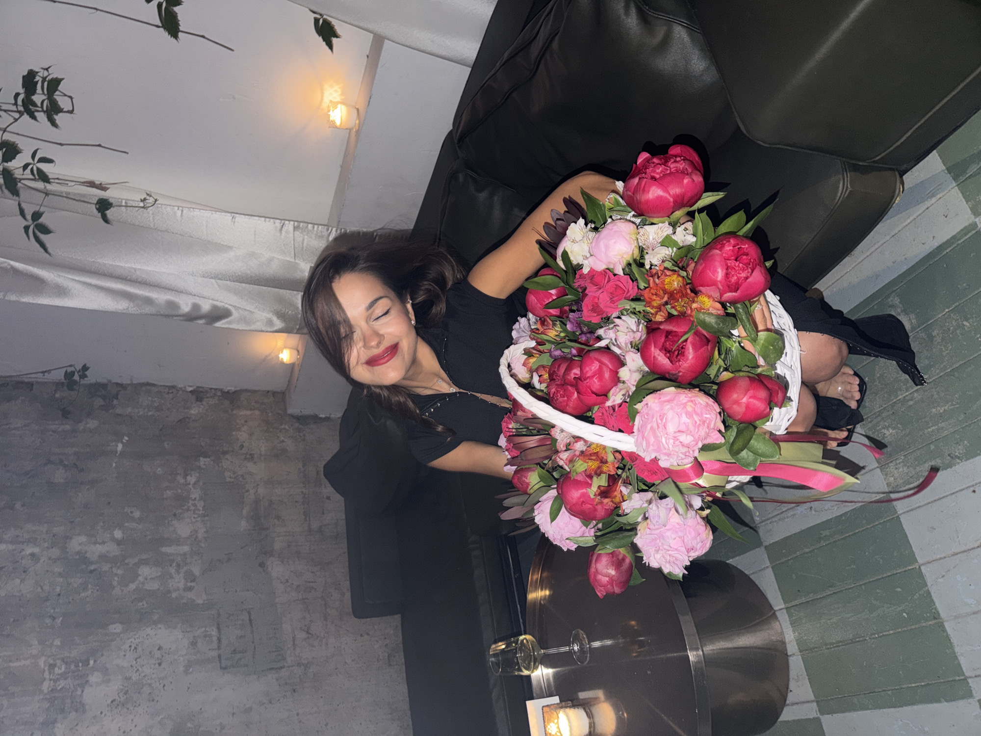 The image shows a woman in her 20s or 30s seated on a dark leather sofa, holding a large white wicker basket brimming with flowers. She has brown hair, wears a dark zippered top, and exhibits a gentle smile with closed eyes and red lipstick. Her feet, visible at the bottom right, have painted toenails and are clad in dark open-toed sandals. The flower basket contains a vibrant assortment of pink and fuchsia peonies, along with orange and light pink smaller blossoms, and is decorated with green and pink ribbons.
To the woman's left, a round metallic coffee table holds a stemmed glass containing a light-colored liquid and a lit candle in a clear holder. Additional lit candles are positioned on a white ledge behind her. The wall to the left is textured and grey, showing signs of distressed plaster, while the wall behind her is lighter with green leafy vines hanging from above. Light-colored curtains are visible on the upper right. The floor consists of alternating dark and light green rectangular tiles. The setting is described as being in Moscow, Russia.