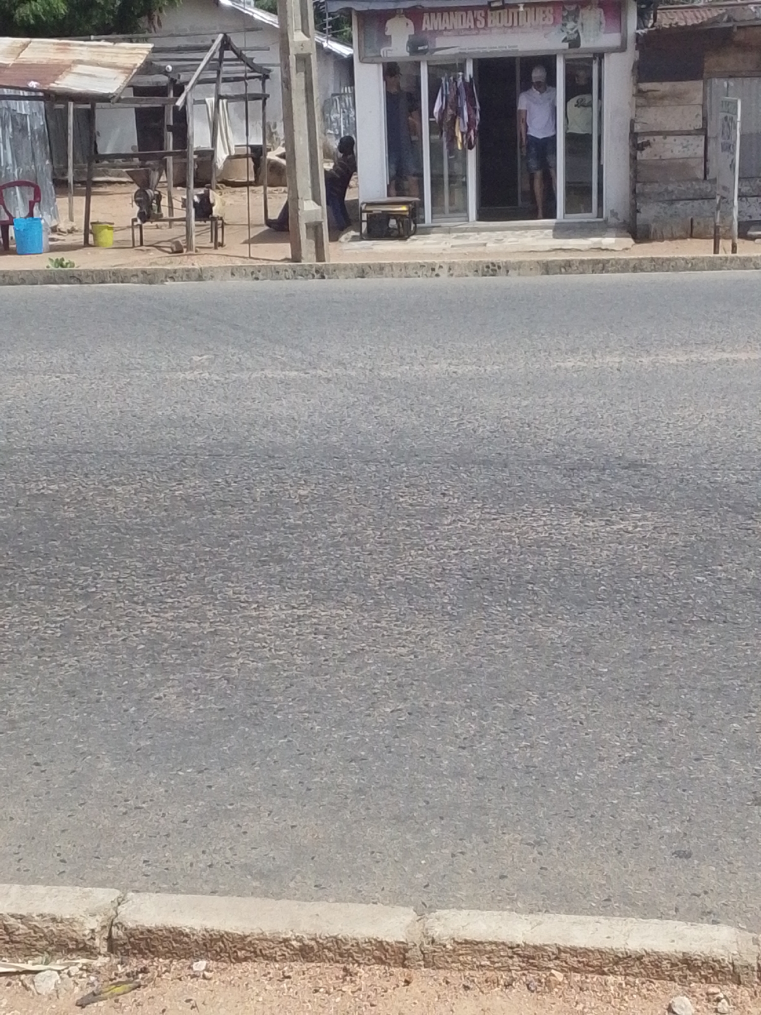 The image shows a street scene in Jalingo, Nigeria. In the foreground, a paved road runs horizontally across the frame. The road has a rough, asphalt texture. A concrete curb separates the road from a dirt shoulder.
In the background, several structures are visible. To the left, there's a lean-to structure with a corrugated metal roof and wooden supports. Inside, a red chair and two buckets, one blue and one yellow, are placed on the ground. Further back, a wooden frame supports what appears to be some sort of machinery, possibly a grinder. A person in a dark shirt and blue jeans is leaning against a pole to the right of this structure, looking up and to the right.
To the right of the lean-to, there is a shop with a sign that reads "AMANDA'S BOUTIQUES". The shop has large glass doors, and inside, clothing racks are visible with garments hanging on them. Two men are standing inside the shop. One man, wearing a white polo shirt and shorts, is standing near the entrance. Another man, wearing a light-colored t-shirt and dark shorts, is standing further inside. There is also a small wooden bench in front of the shop. Adjacent to the shop, there are wooden walls forming part of another structure. A concrete utility pole stands to the right of the shop.
The scene appears to be outdoors, likely during the day, given the shadows. The overall impression is that of a commercial area in a town or city, with businesses and street life. The presence of people suggests ongoing activity.