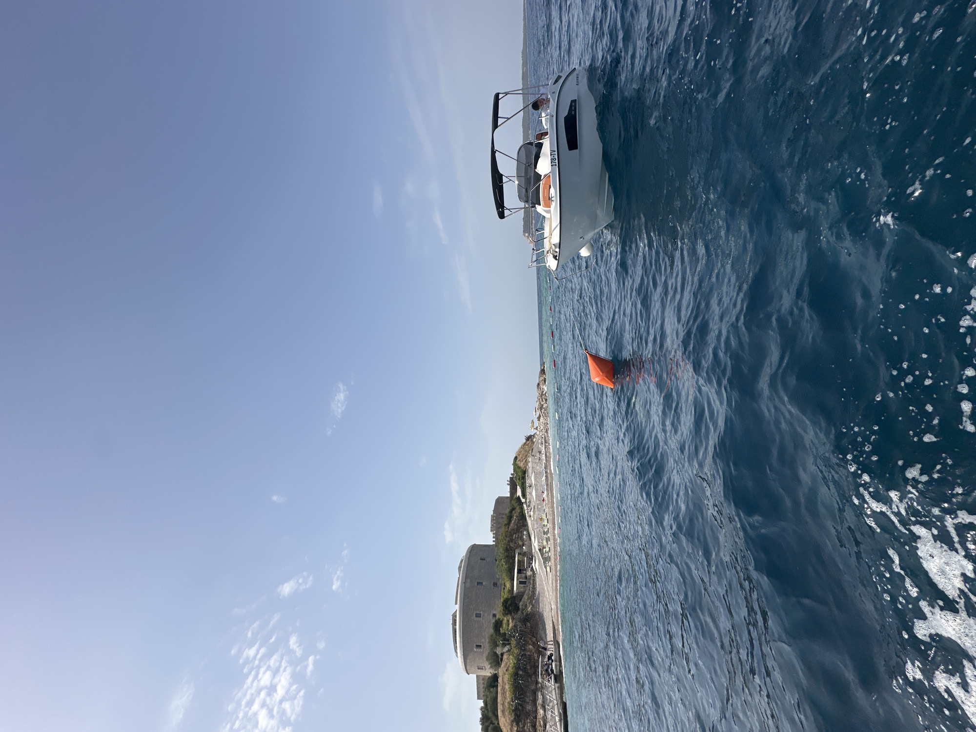 A wide-angle, low-angle shot captures a boat on a clear, blue sea under a bright, cloud-dusted sky. In the background, a large, circular, stone fortress stands on a small island, accessible by a walkway with a few figures visible. The island also features a beach lined with lounge chairs and umbrellas. The boat, white with a black canopy and a registration plate reading "178-TV", is positioned in the foreground, to the right of center, with a person seated inside. An orange buoy is visible in the water between the boat and the island. The water exhibits moderate waves and some white foam. The scene appears to be a view of Forte Mare, a historic fortification located in Herceg Novi, Montenegro, situated on the Luštica peninsula.