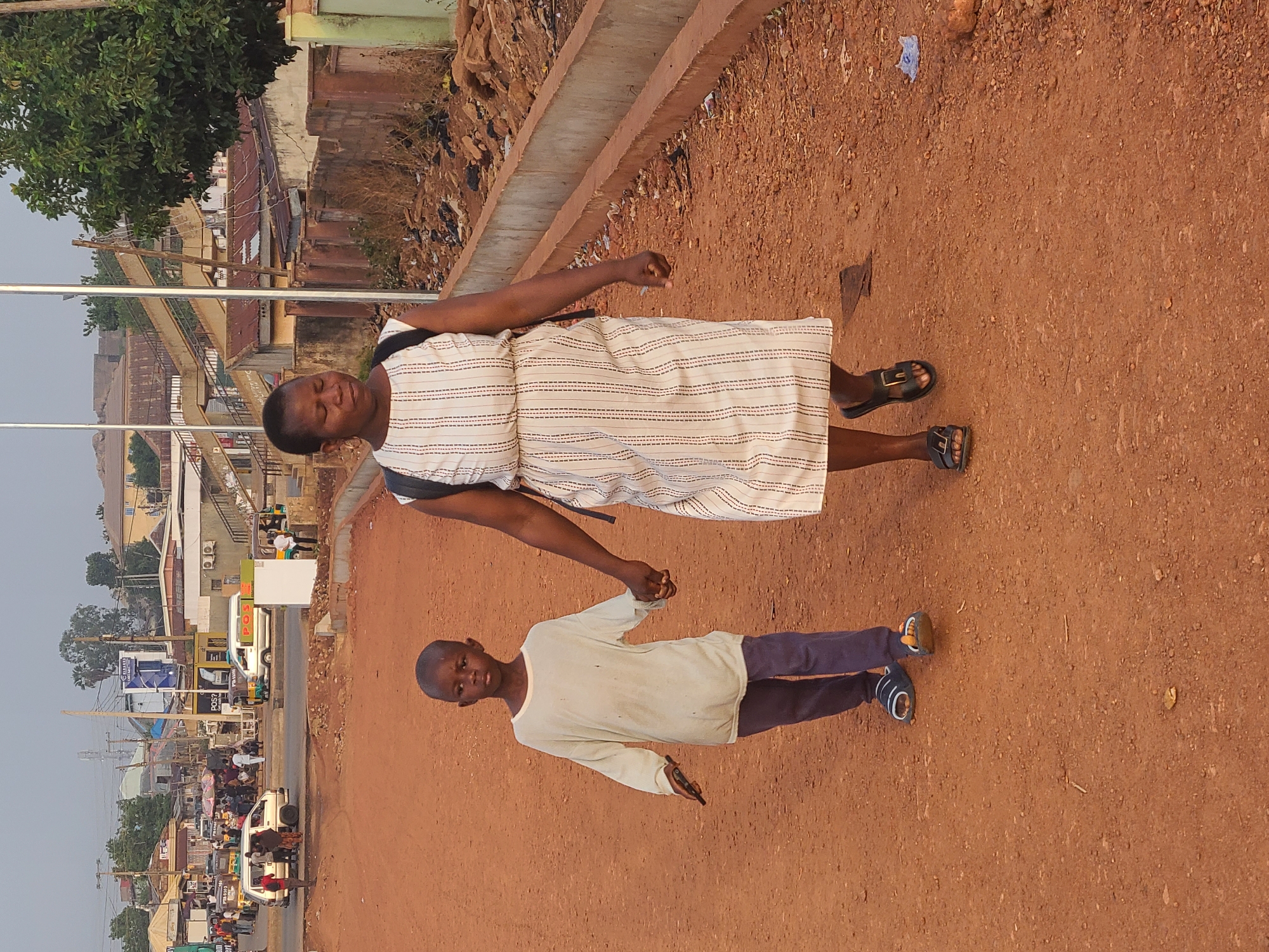 A medium outdoor shot shows a woman and a boy walking together along a dirt path in Jos, Nigeria. The woman, wearing a white dress with dark vertical stripes, has dark hair, dark skin, and sandals. The boy, wearing a long-sleeved white shirt and dark blue pants, has dark skin, a shaved head, and blue and brown shoes. They are holding hands, walking towards the right side of the frame. Behind them, a concrete curb separates the dirt path from a busy street with cars, buildings, and billboards. The sky is overcast.