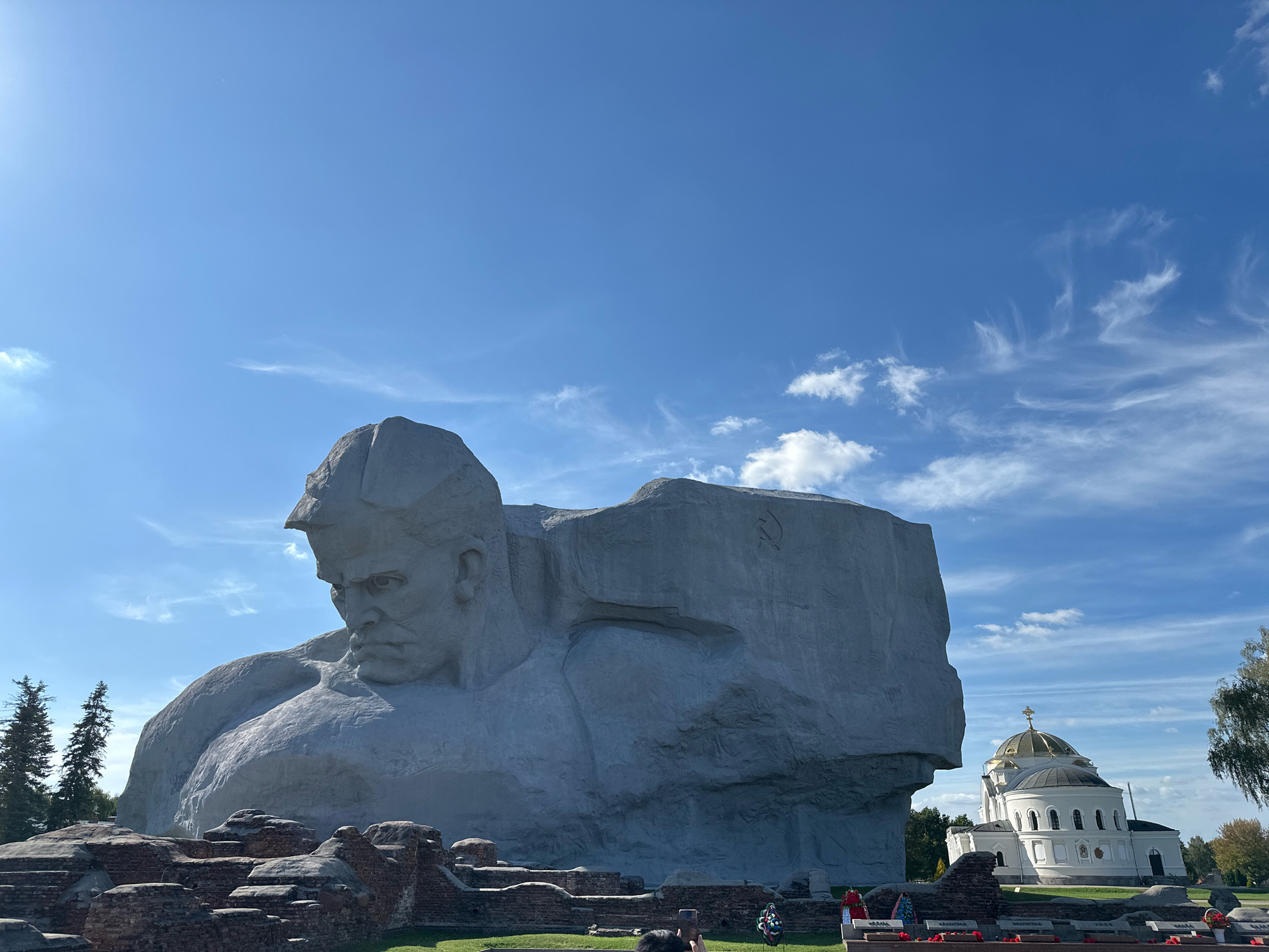 The image shows the monumental "Brest Fortress" monument in Brest, Belarus. The monument, a colossal grey sculpture of a soldier's head and torso, dominates the frame. To the right of the monument is a white church with a golden dome, characteristic of Orthodox Christian architecture. In the foreground, there are remnants of ruined brick walls and a grassy area with several memorial plaques and floral tributes. The sky is a bright blue with scattered white clouds. A few trees are visible on the left and right sides of the frame, and the sun is shining brightly, casting light on the scene. In the lower portion of the image, a person is partially visible, holding up a phone, likely to take a photograph or video of the monument.