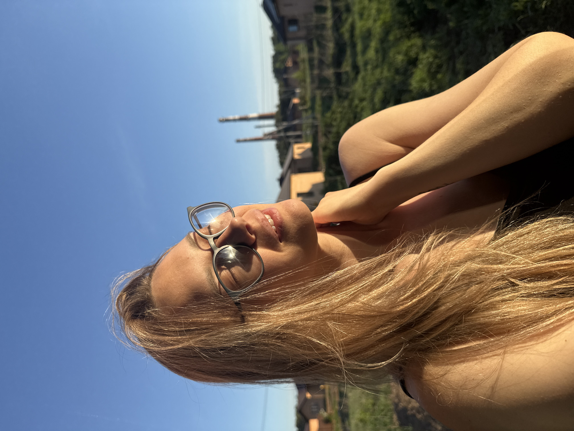 A young woman with blonde hair and glasses is looking up towards the sky in an outdoor setting. The background indicates the industrial landscape of Chaikovsky, Russia, evidenced by the presence of chimneys and factory structures in the distance. The foreground features the woman's face, hair, and upper body, suggesting she might be relaxing or sunbathing. The environment appears sunny, with a clear blue sky visible above the urban landscape.