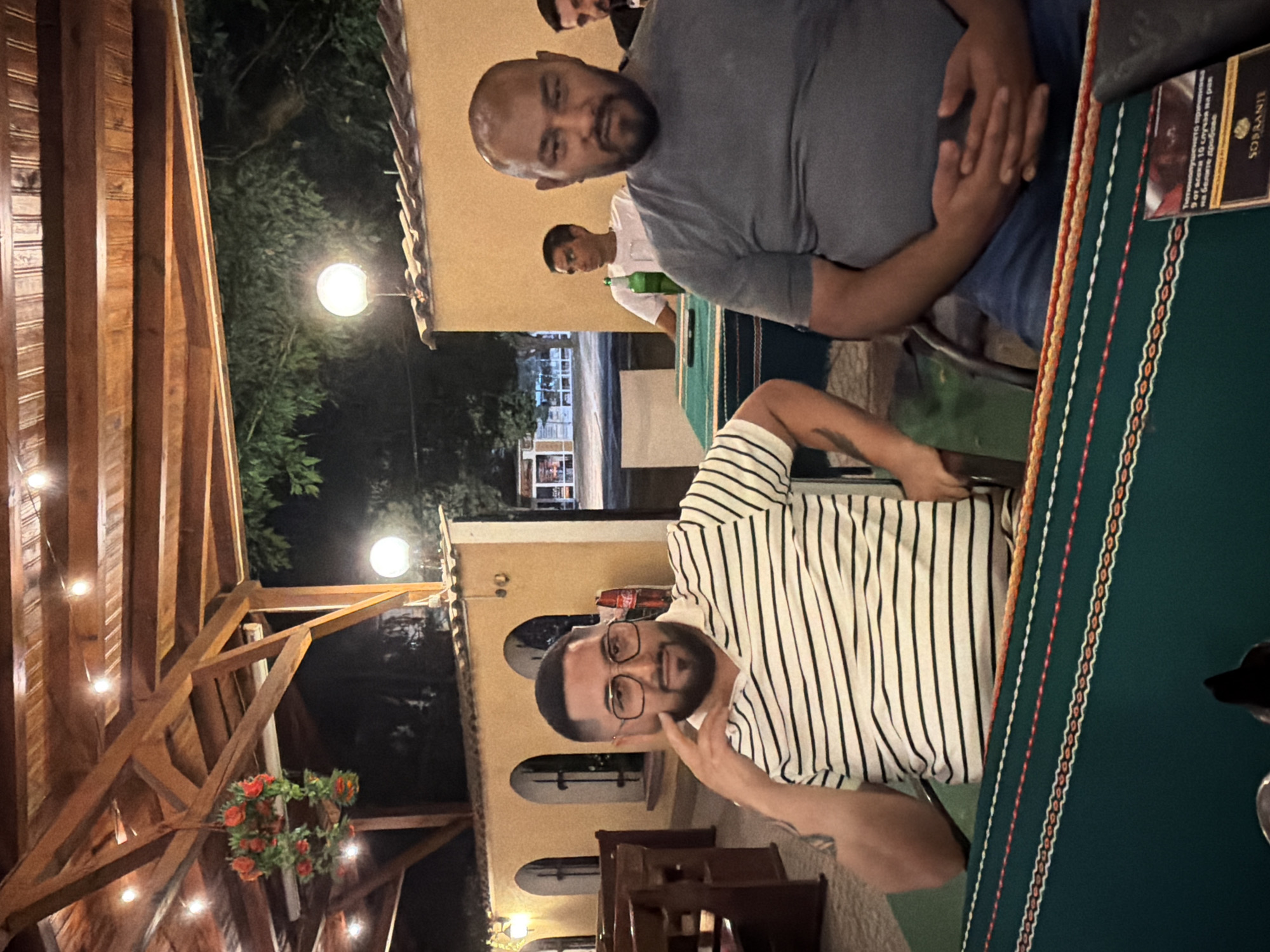 The image depicts an outdoor dining scene at night, possibly a restaurant or a tavern, in the city of Levski, Bulgaria. The setting features a wooden-beamed covered patio illuminated by string lights and a larger globe light. In the foreground, two men are seated at a table covered with a green tablecloth featuring decorative trim. The man on the left, wearing a black and white striped shirt and glasses, is looking towards the camera with his hand near his face. The man on the right, with a shaved head and beard, is wearing a grey t-shirt and blue jeans and is looking towards the camera with his hands clasped. In the background, another man in a white t-shirt is visible, and behind him, a street scene with buildings and a Coca-Cola sign is discernible. The overall atmosphere appears casual and social, suggesting a meal or gathering.