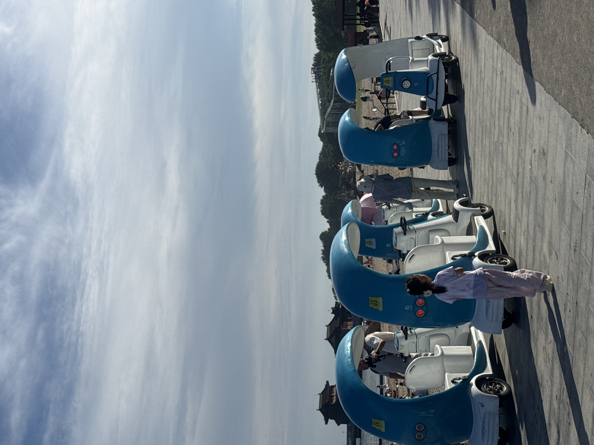 The image captures a scene on a paved walkway by a beach in Qinhuangdao, China, likely near a coastal attraction. In the foreground, a young girl in a light purple shirt and pants walks away from the camera, casting a long shadow. She is positioned between several whimsical, blue and white mini-vehicles, some resembling stylized waves or moons, designed for personal transport. One of these vehicles has the number "03" on it. Behind her, an adult male in a light-colored shirt and a person with a striped shirt and a hat are standing near another blue mini-vehicle. Further in the background, other people are visible on the beach and near structures that appear to be traditional Chinese architecture, including a dark-colored building with a tiered roof. The sky is partly cloudy, with a mix of blue sky and white clouds. The overall atmosphere suggests a leisure or recreational area.