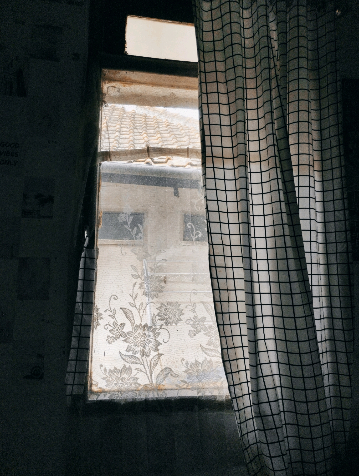 The image shows a window with a patterned curtain and a view of tiled roofs through the glass. The window is divided into sections, with a decorative floral pattern on the lower part of the glass. A curtain with a black and white grid pattern is partially drawn across the window on the right side. The left side of the image shows a dark wall with some faint text. The overall impression is one of a domestic interior, possibly in an older building, with natural light filtering in. The location is identified as Cimenyan, Indonesia, suggesting this scene is within a residential or urban area in that region.