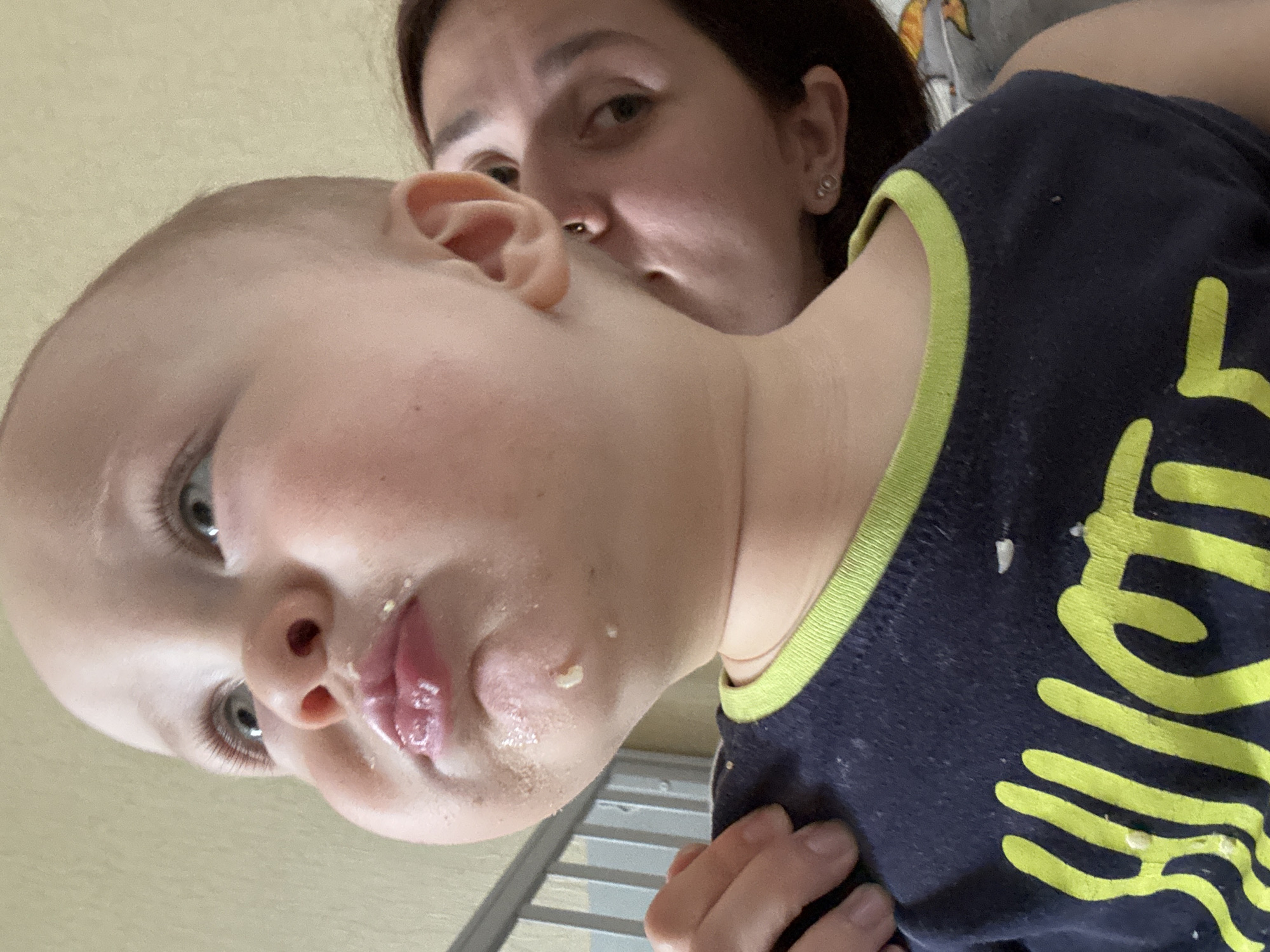 A close-up, over-the-shoulder shot features a baby with chubby cheeks, looking upwards and to the left. Food particles are visible on the baby's face and shirt, which is dark blue with a neon green trim and a yellow graphic. A woman's face is partially visible on the right side of the frame, looking towards the baby. The background is a plain, light-colored wall. The overall scene appears to be indoors, possibly in a home environment. The city of Naberezhnye Chelny, Russia is not depicted in this image.