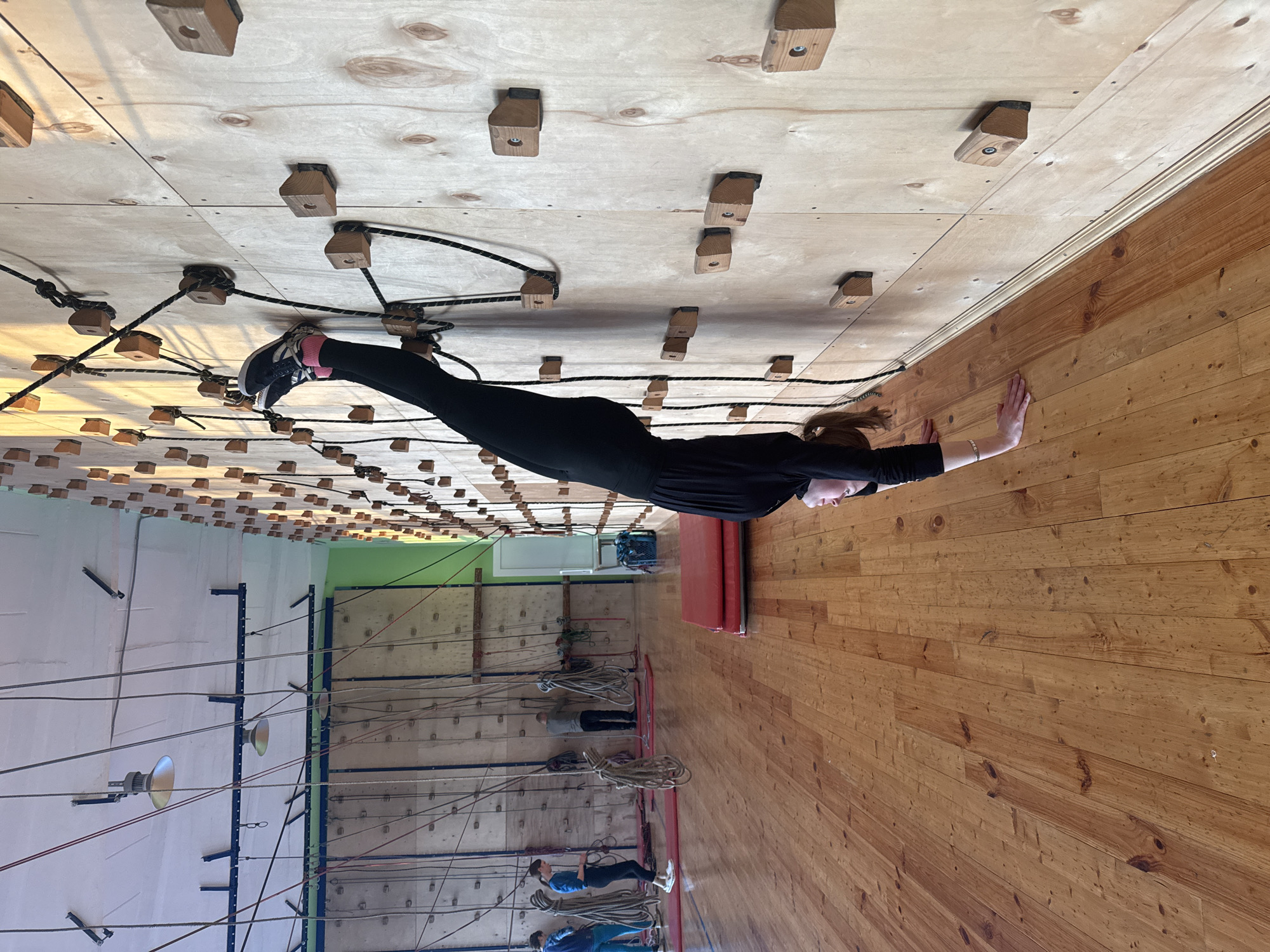 The image shows a woman suspended horizontally between two climbing walls in what appears to be an indoor climbing facility. She is wearing black clothing and sneakers, with her feet positioned on a climbing wall with wooden holds connected by ropes. Her hands are pressing against the opposite wall, which is made of wooden planks.
The climbing facility has additional climbing walls with safety ropes and belay systems visible in the background. The overall environment suggests a recreational or training space for climbing activities. The location is Chaikovsky, Russia, based on given location data.