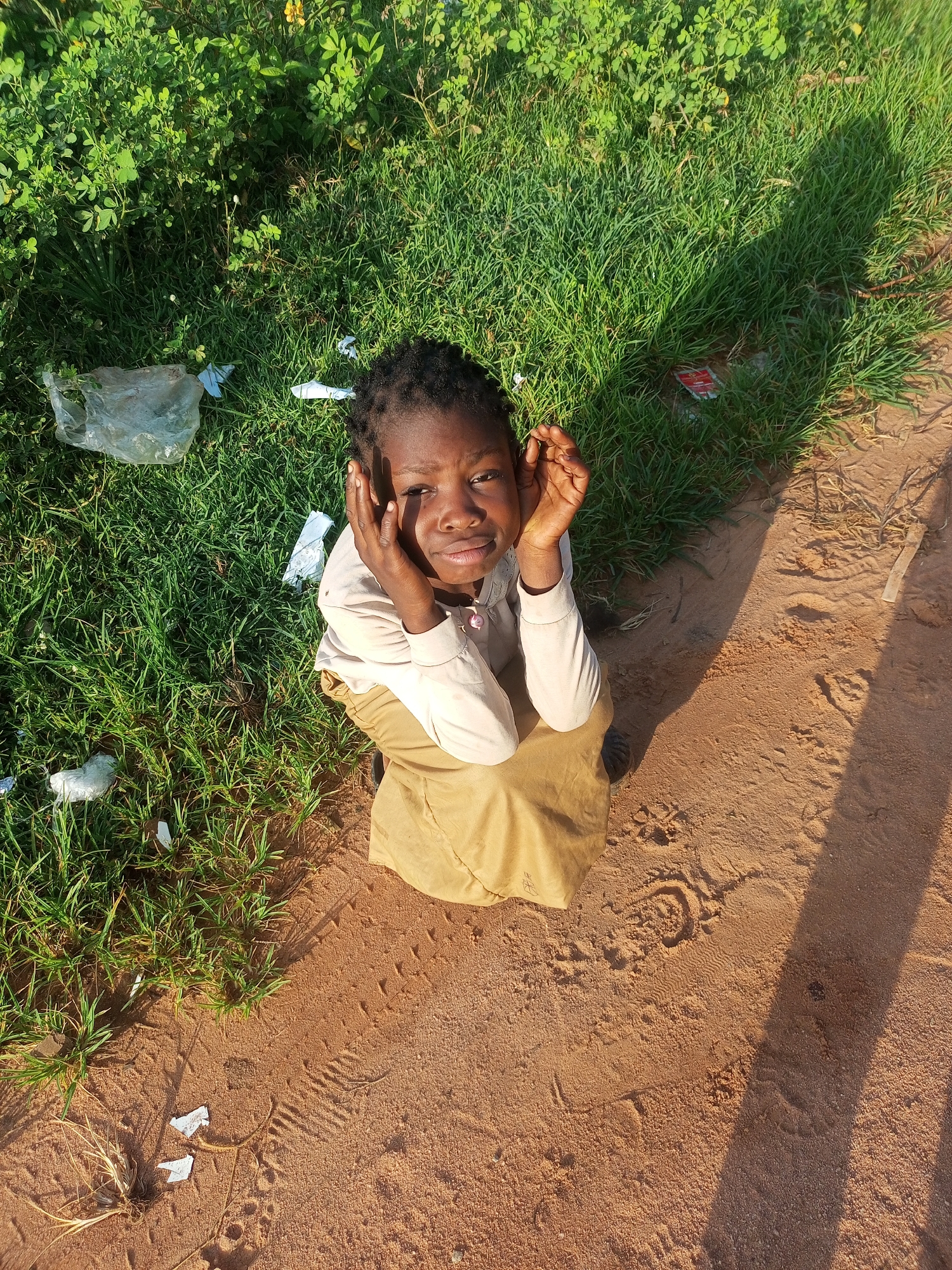 In Tempure, Nigeria, a young Black girl sits on a dirt path next to a grassy area. She is wearing a light-colored long-sleeved shirt and a tan skirt. Her hands are raised to her face, and she is looking directly at the camera with a thoughtful expression. The ground is covered in loose dirt and some sparse vegetation, with a few pieces of white plastic trash scattered on the grass. Sunlight casts a shadow across the dirt path, indicating it is a sunny day.