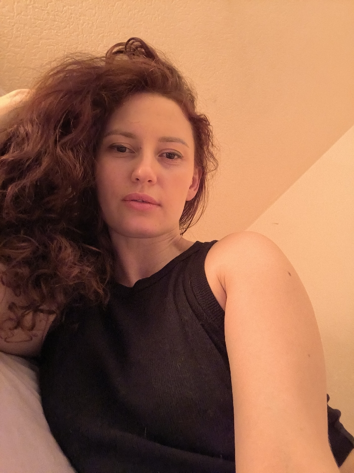 The image displays an indoor, low-angle portrait of an adult female. She has long, wavy, reddish-brown hair, a fair complexion, and a neutral facial expression with slightly parted lips, looking directly into the camera. She is wearing a dark, sleeveless garment, possibly a tank top or sweater, with a visible ribbed texture on the shoulder strap. Her left arm is partially visible on the left side, behind her head/shoulder area, which rests on a white pillow or bedding. Her right arm extends into the lower right of the frame, showing the bicep and forearm. The background consists of a plain, light peach-colored wall, exhibiting a subtle texture. A diagonal line in the upper right suggests an angled ceiling or wall corner. No identifying features within the image indicate the specific location in Saint Petersburg, Russia, or any other city.