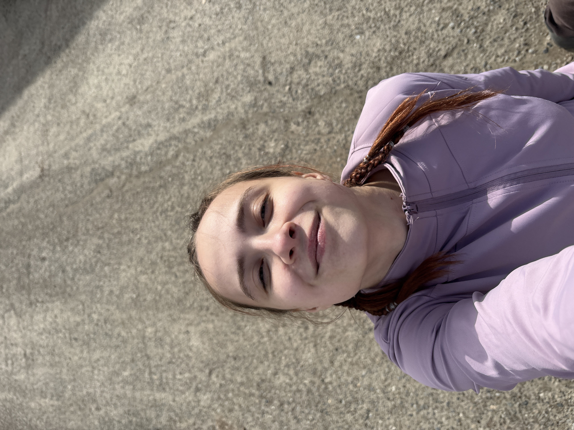 A young woman is captured in a close-up, upward-angled shot outdoors in Alnashi, Russia. She has a light complexion, dark eyebrows, and brown hair with visible reddish or auburn braided sections, some loose strands framing her face. She is wearing a light purple, long-sleeved, collared zip-up jacket, which is partially visible. Her head is tilted slightly, and she looks directly at the camera with a relaxed expression, a subtle smile on her lips, and her eyes partially closed, possibly squinting due to bright sunlight.
The background primarily consists of a textured, light gray surface, likely asphalt or concrete, suggesting a paved outdoor area. The lighting is bright and natural, indicating daytime, with subtle shadows cast on her face and jacket, and more pronounced shadows visible on the ground. No visible text is present in the image.