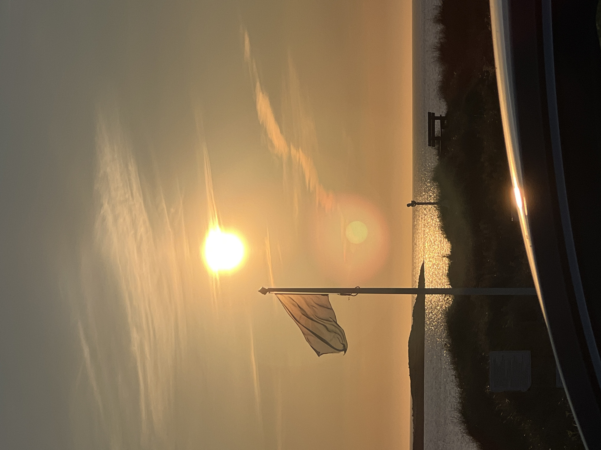 The image depicts a sunset scene at the coast in Lunniaghbeg, Ireland. The sun, a bright orb, is positioned high in the sky, casting a warm, golden light across the scene and reflecting intensely on the water. A white flag flutters from a flagpole on a grassy dune, positioned to the left of the frame. In the middle ground, there is a dark silhouette of land and sea, with a small island visible in the distance. Further along the coastline, a wooden bench and a dark silhouette of what appears to be a person or a structure are situated on the dune. The foreground is partially obscured by the dark, curved surface of a vehicle. The sky is a gradient of warm tones, transitioning from bright yellow near the sun to a softer orange and then a dusky grey at the top of the frame, with wisps of clouds streaked across it. The overall atmosphere is serene and captures the beauty of a coastal sunset.